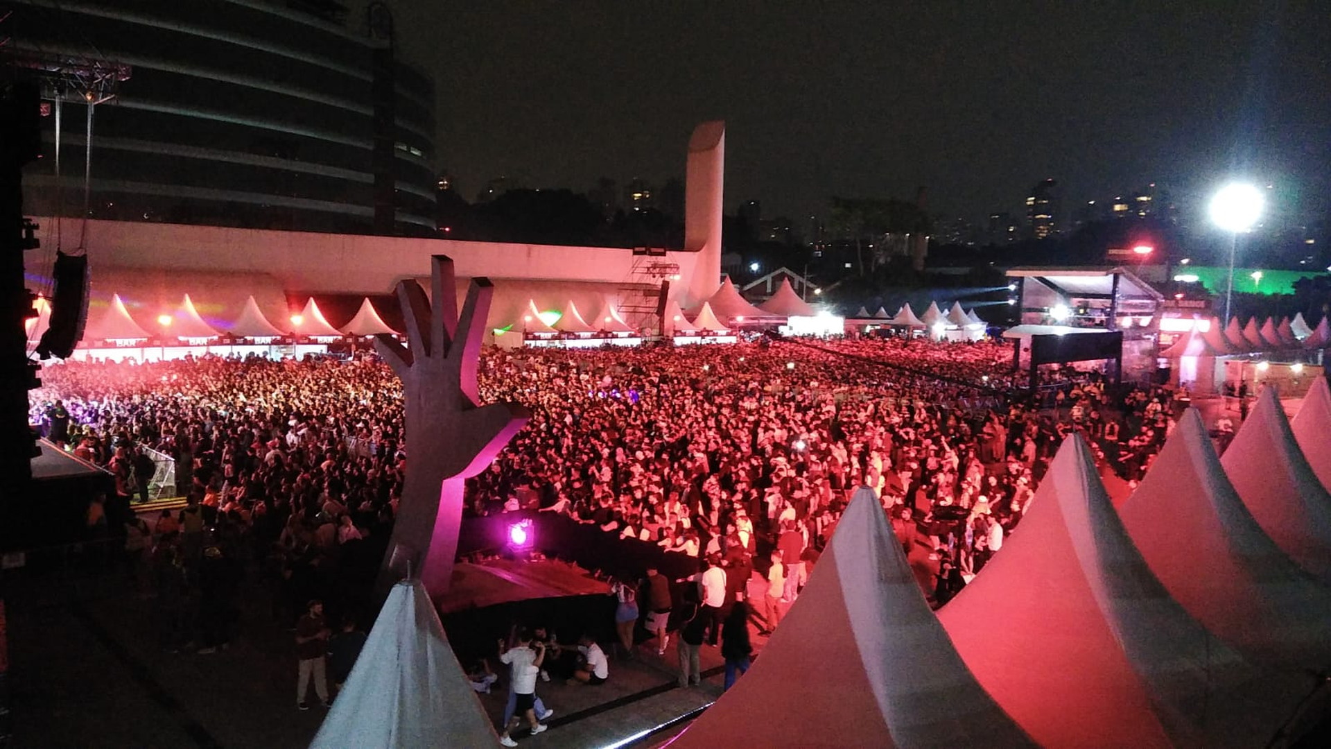Vista nocturna de una multitud ante carpas blancas y estructuras iluminadas en rojo, con una escultura angular en primer plano