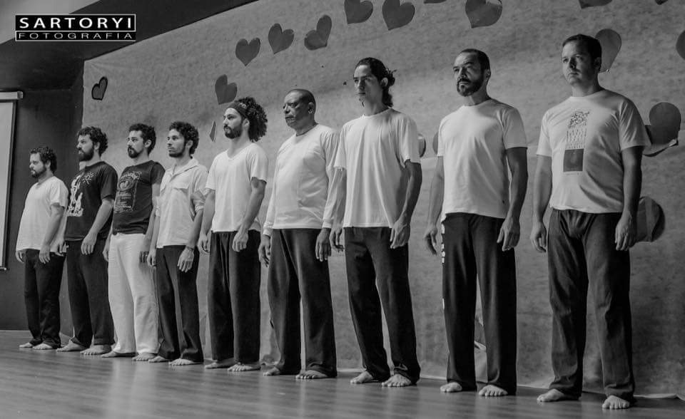 Elenco masculino alinhado em um palco durante registro de trabalho cênico em preto e branco