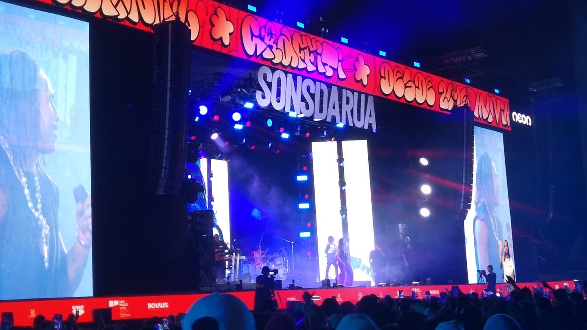 Escenario iluminado con pantallas que muestran a una persona y público ante el espectáculo, con teléfonos móviles en alto