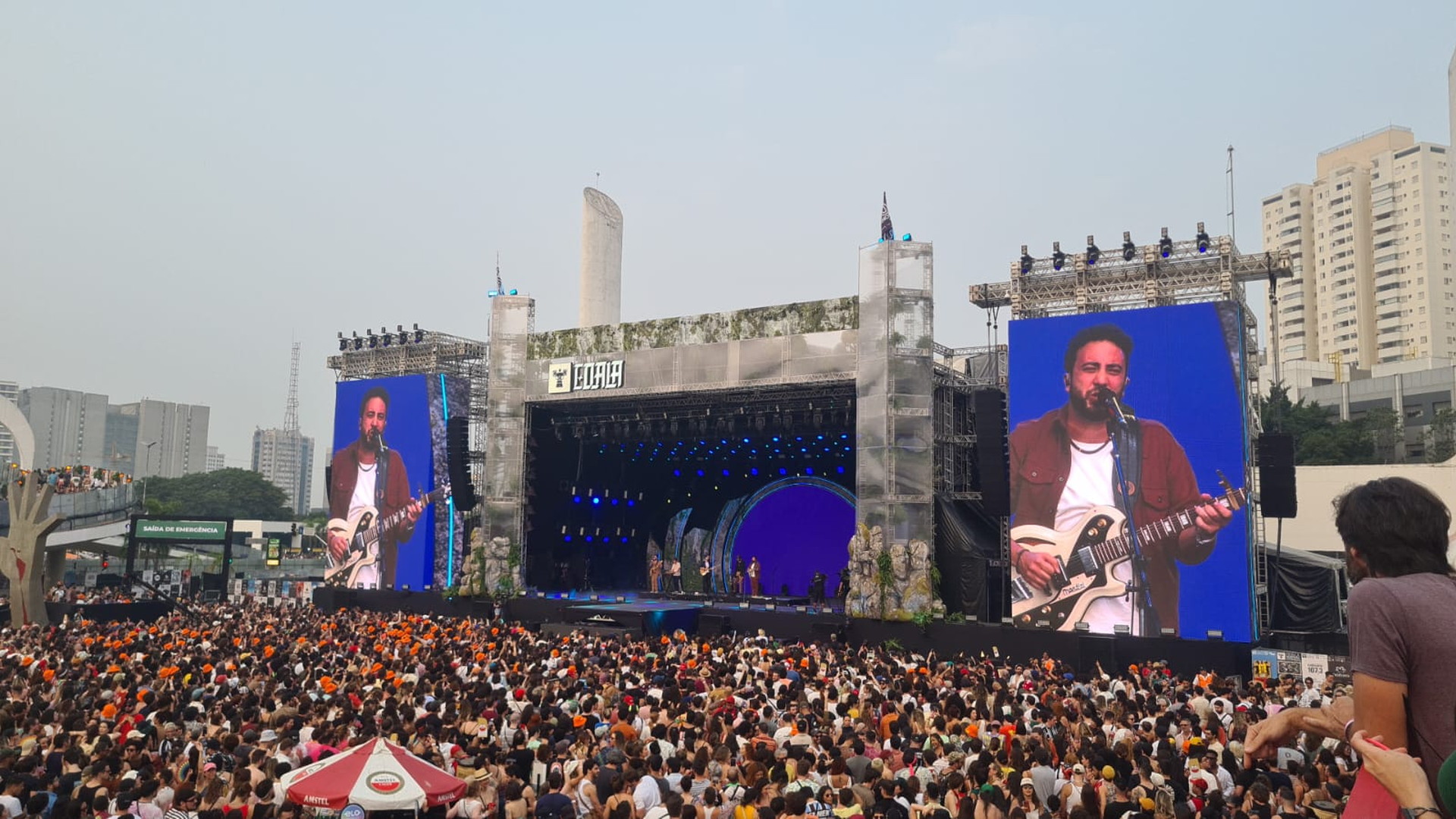 Gran público ante un escenario exterior iluminado en azul, con pantallas que muestran a un músico tocando la guitarra y edificios al fondo