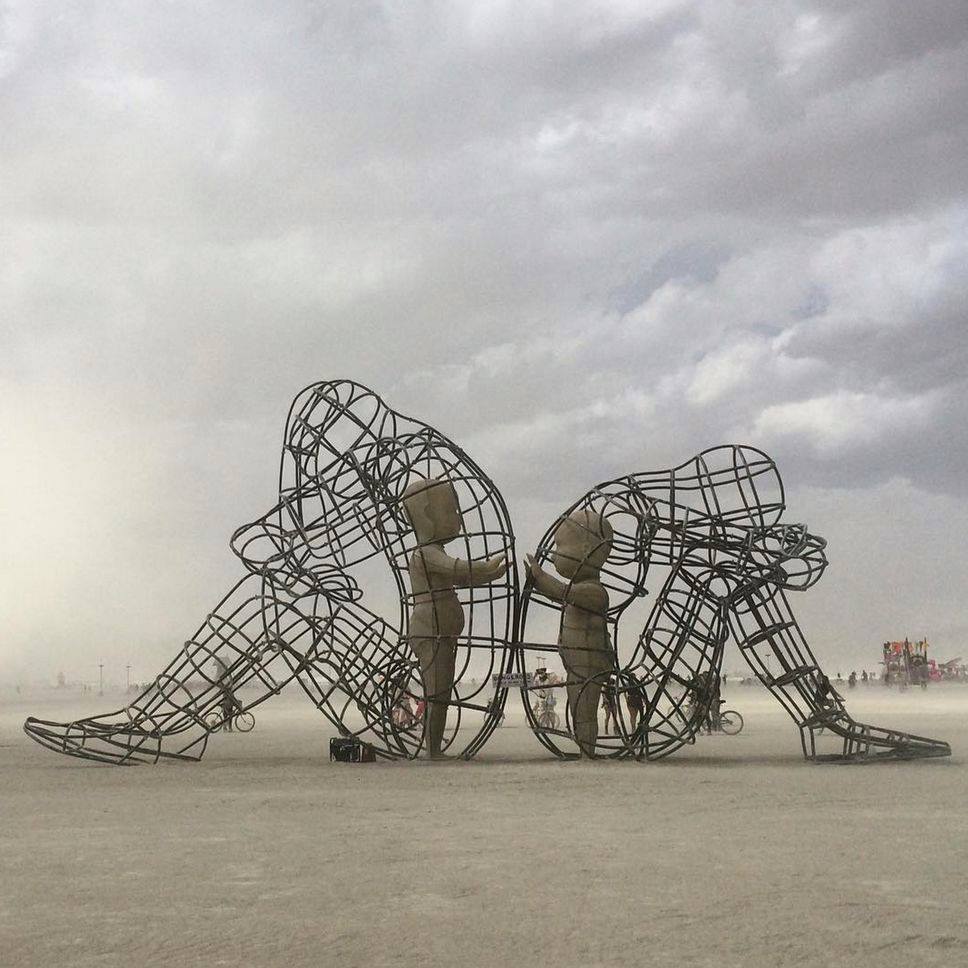 Estruturas artísticas de grandes figuras humanas sentadas, fotografadas em paisagem aberta sob céu nublado