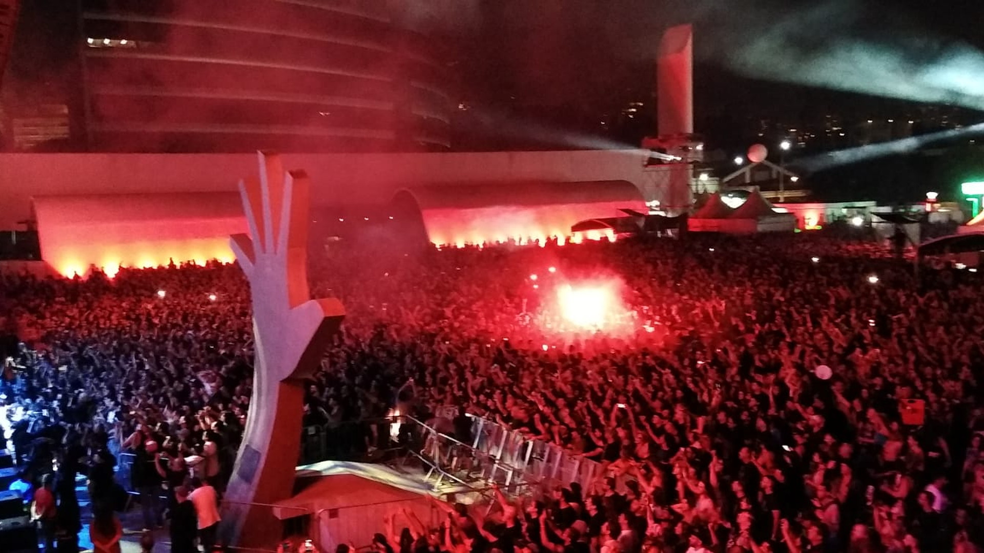 Multitud de noche con los brazos en alto, bañada por luz roja, ante una gran estructura blanca y haces de luz