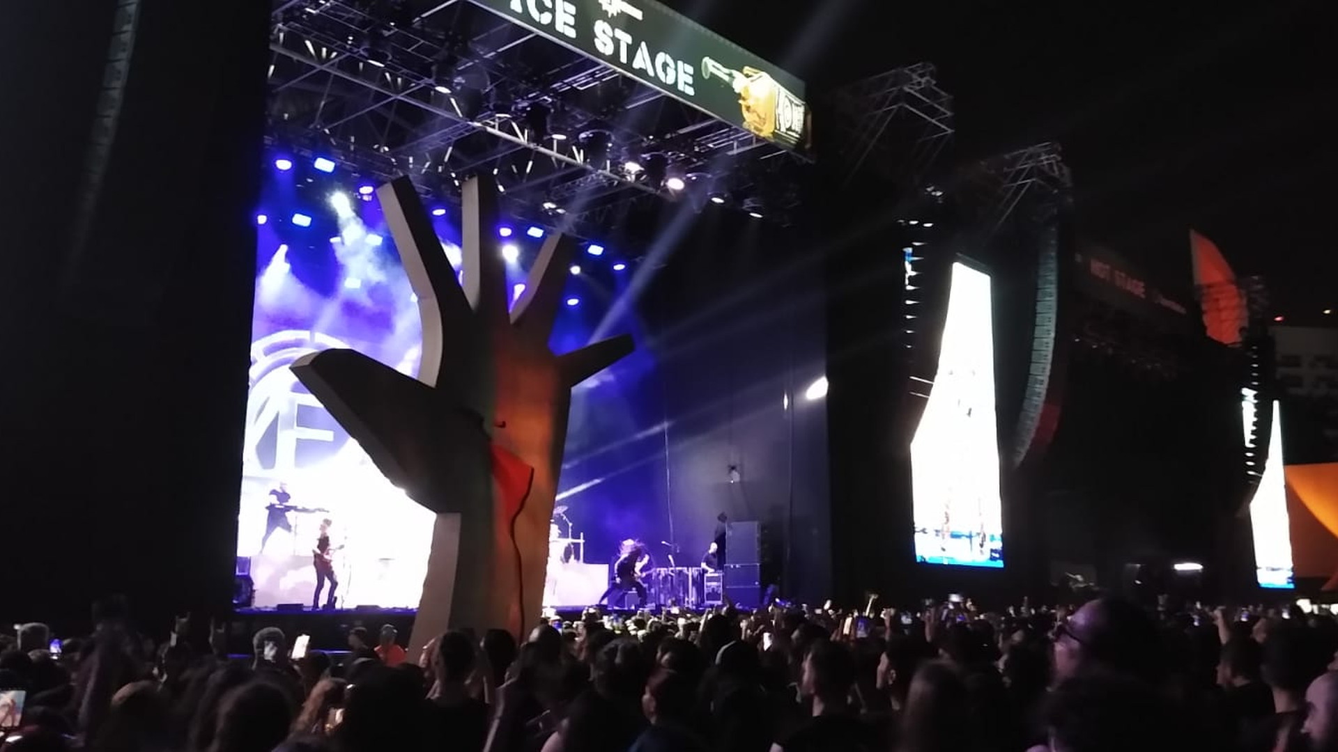 Escenario iluminado de noche ante una gran multitud, con teléfonos móviles en alto, paneles de video y una escultura escenográfica en primer plano