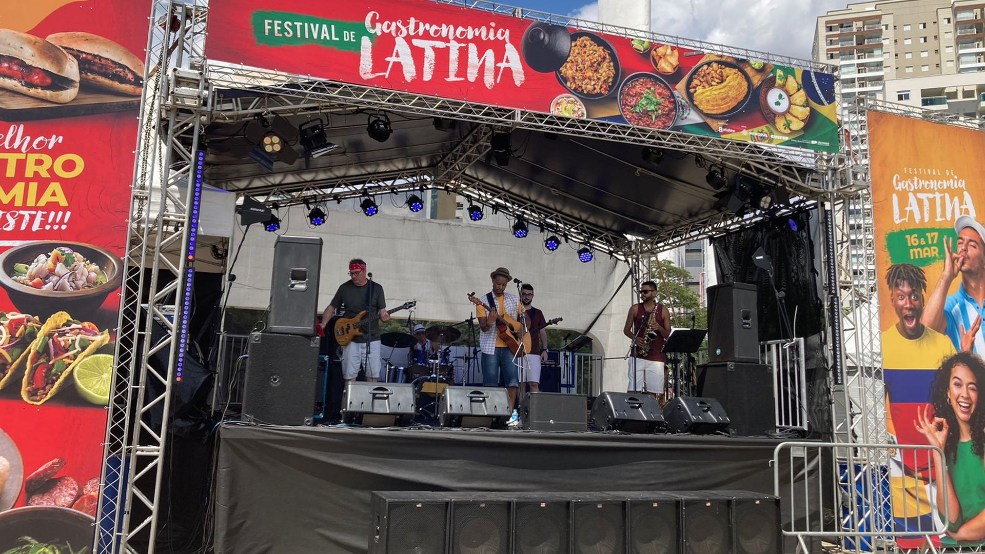 Escenario exterior con cuatro músicos tocando ante carteles del Festival de Gastronomia Latina, con altavoces y edificios al fondo