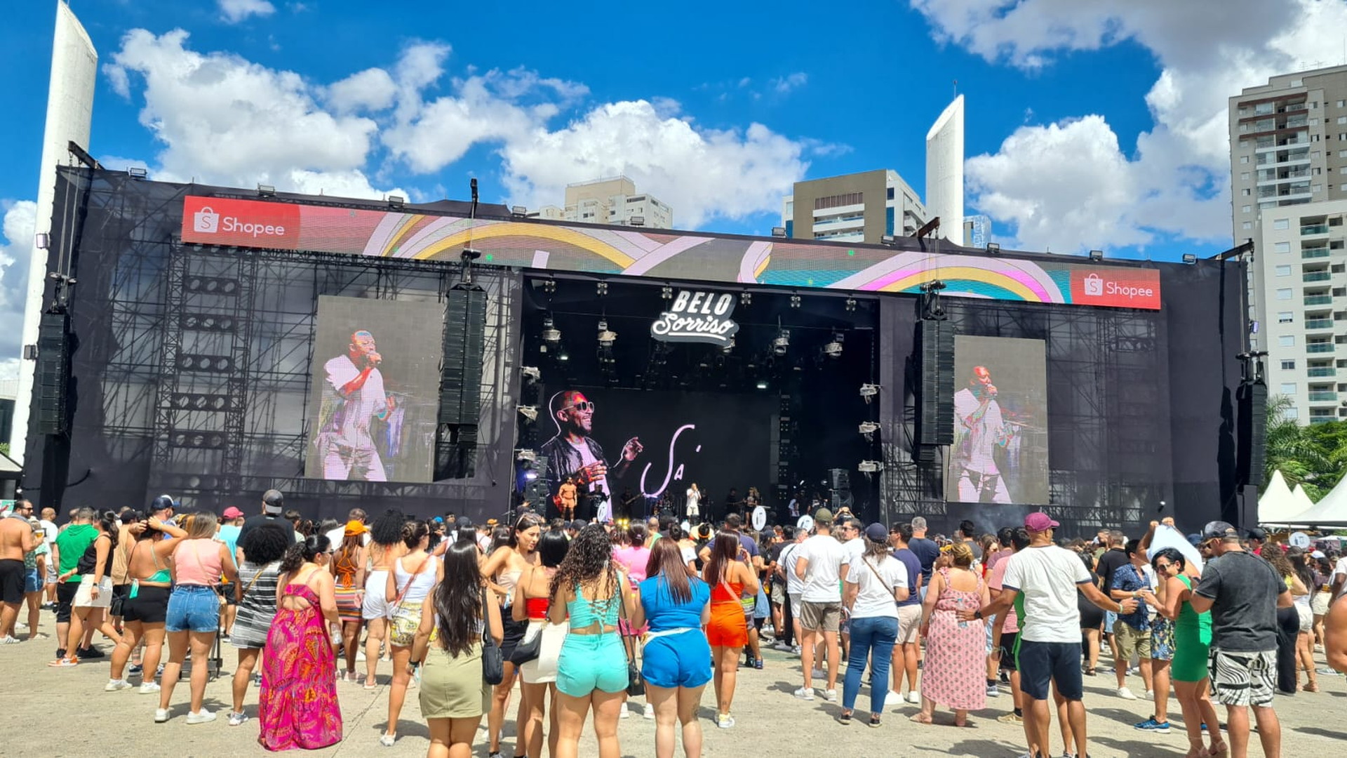 Multitud ante un gran escenario exterior con pantallas, estructuras de sonido y edificios altos al fondo