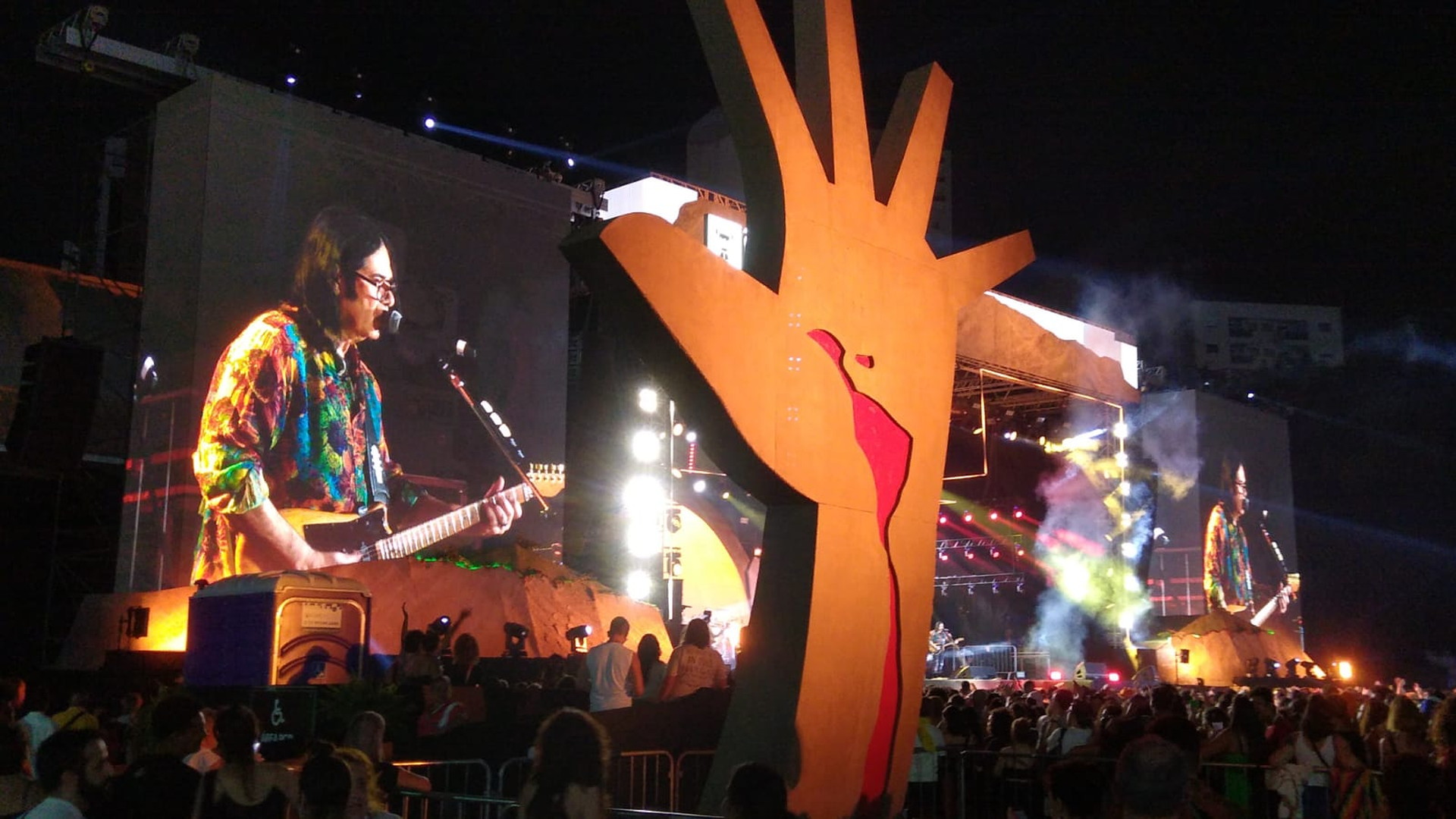 Público ante un escenario nocturno con un cantante y guitarrista en las pantallas y una gran escultura naranja en primer plano