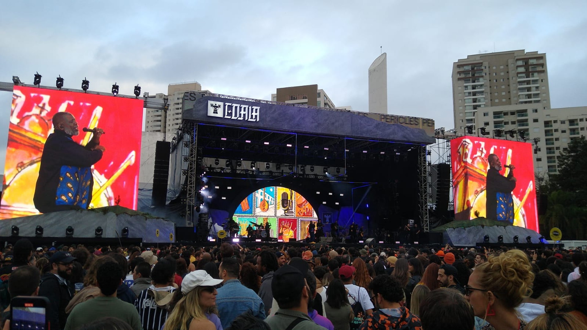 Escenario de concierto al aire libre con un artista al micrófono en las pantallas y multitud ante la estructura