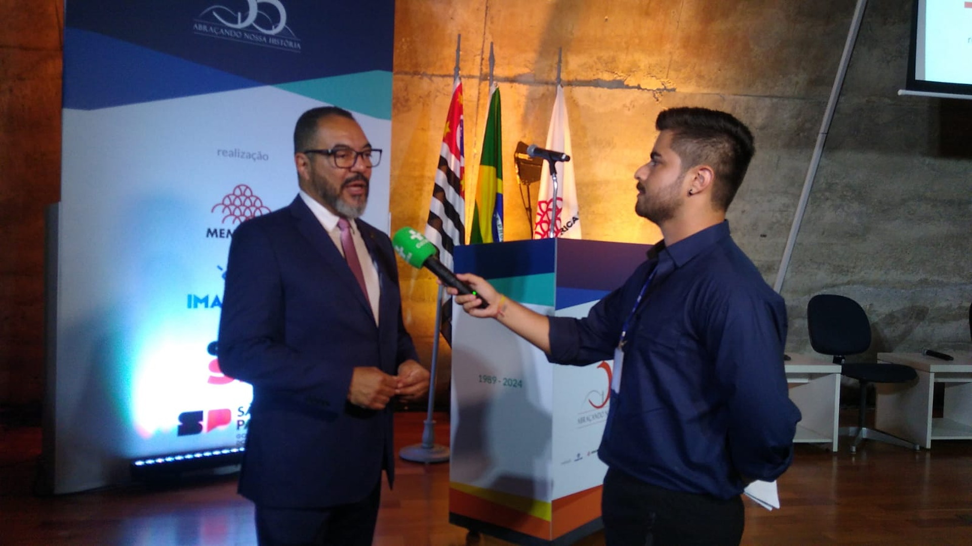 João Carlos Corrêa y un entrevistador conversan en un escenario con una pantalla y banderas al fondo