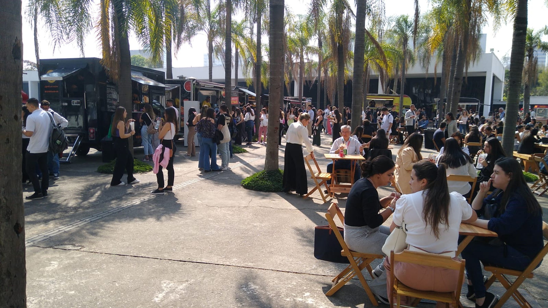 Personas hacen fila en puestos móviles de comida y conversan sentadas en mesas bajo palmeras