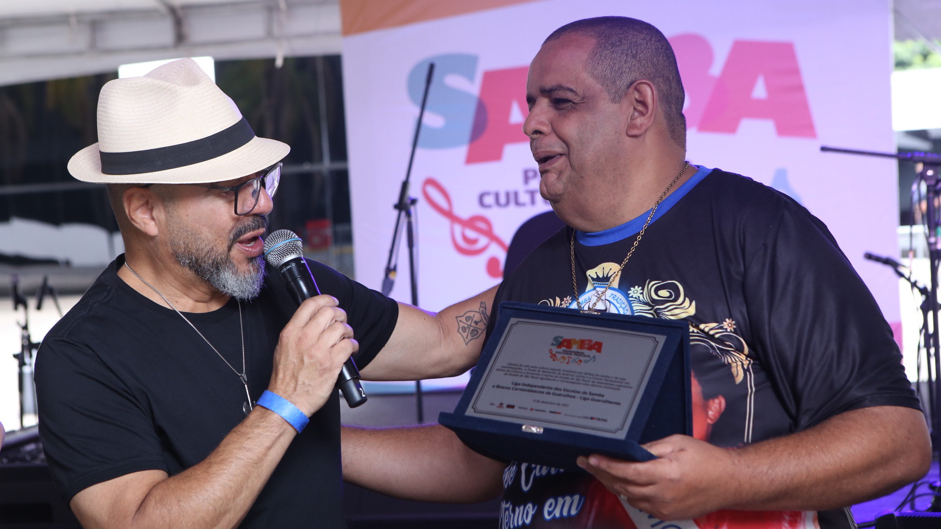 João Carlos Corrêa entrega una placa de reconocimiento a un participante en un escenario de la programación cultural