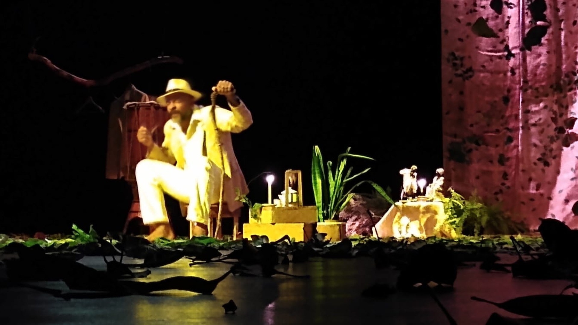 Artista con vestuario claro actúa en un escenario teatral rodeado de objetos escénicos e iluminación dramática