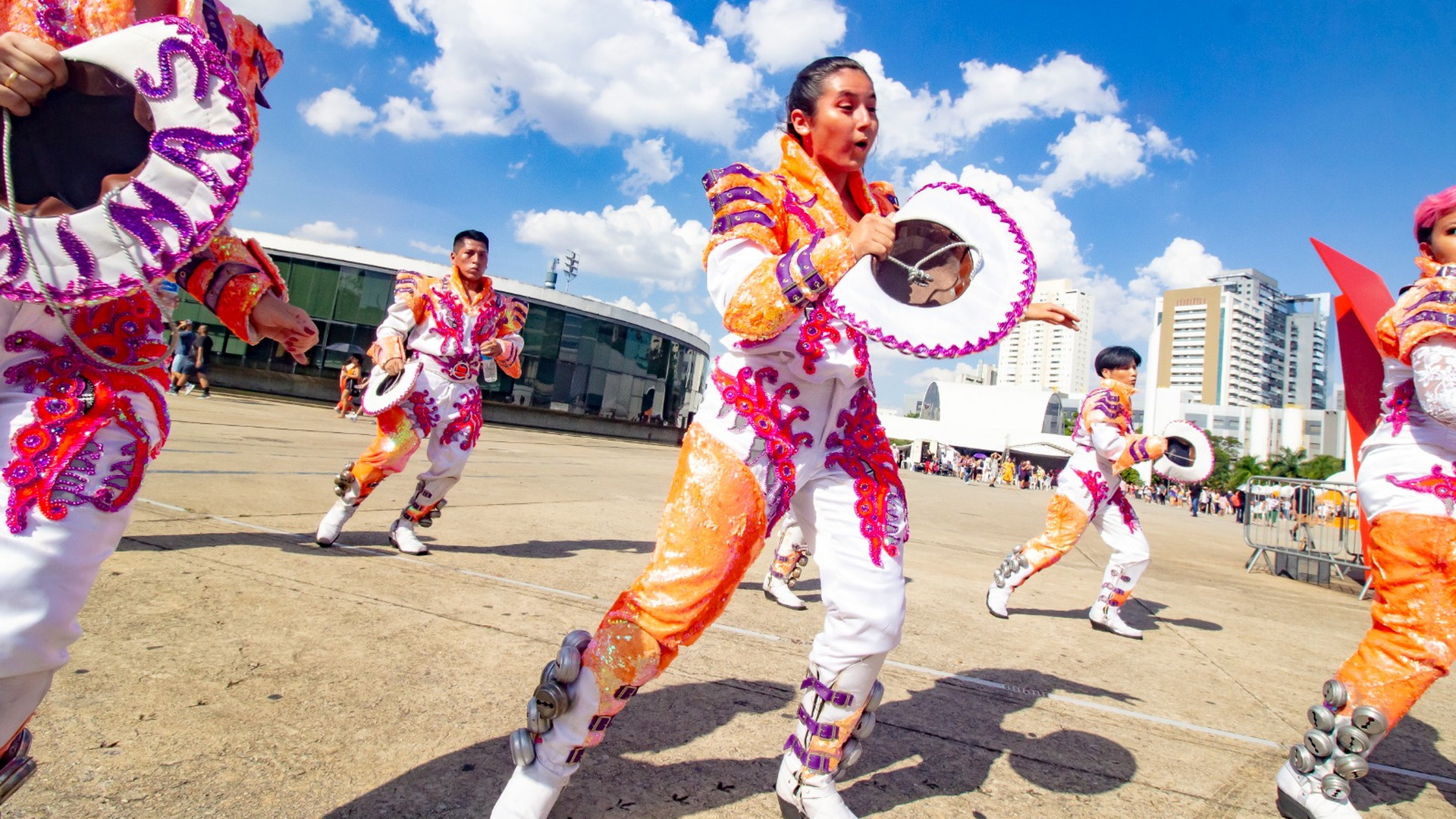 Grupo de bailarines presenta una coreografía con trajes coloridos ante la escultura Mão y el conjunto arquitectónico