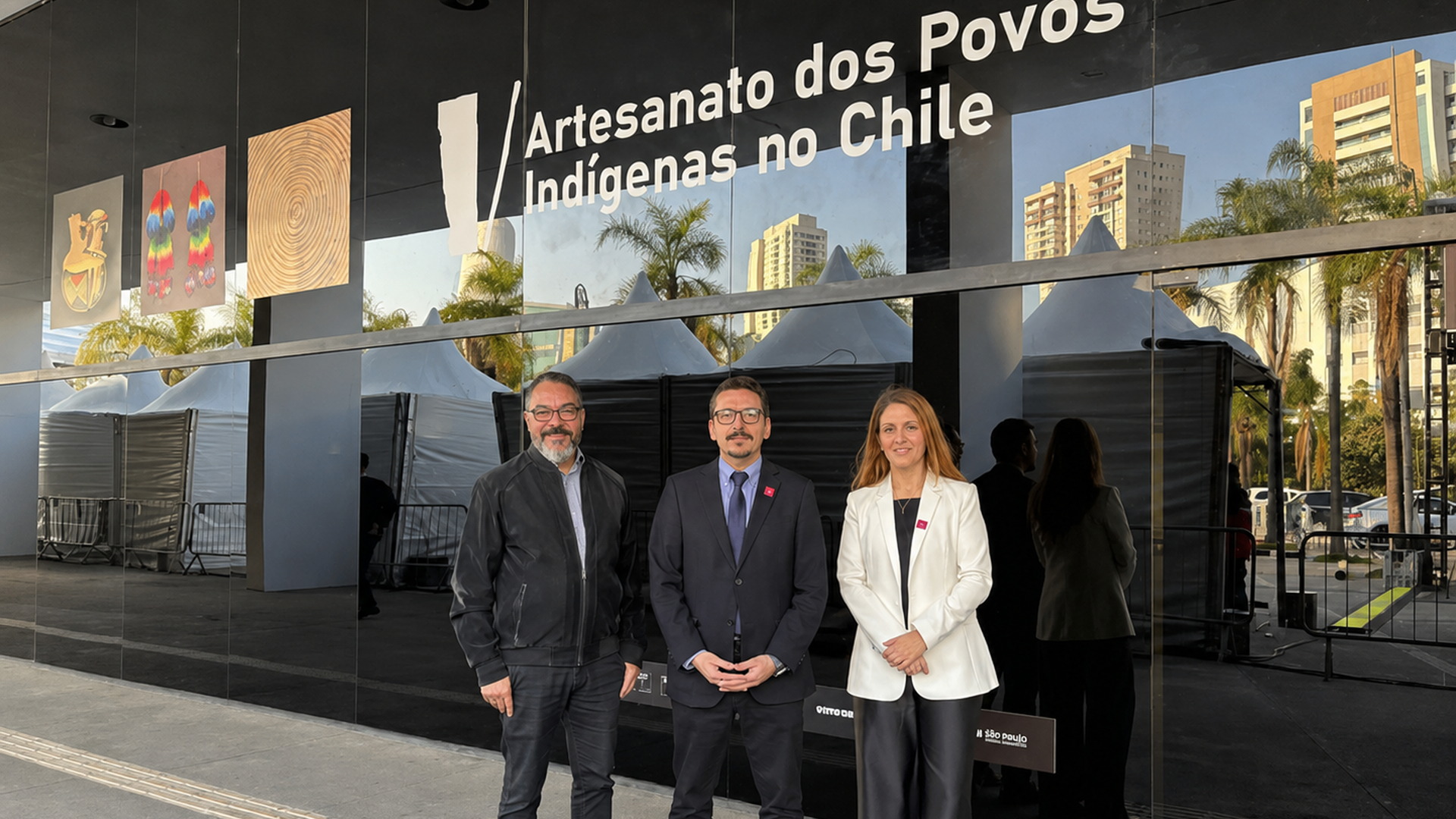 João Carlos Corrêa y dos invitados posan ante la entrada de la exposición Artesanato dos Povos Indígenas no Chile