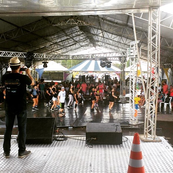 João Carlos Corrêa acompanha do palco uma atividade de dança com participantes e público em espaço coberto