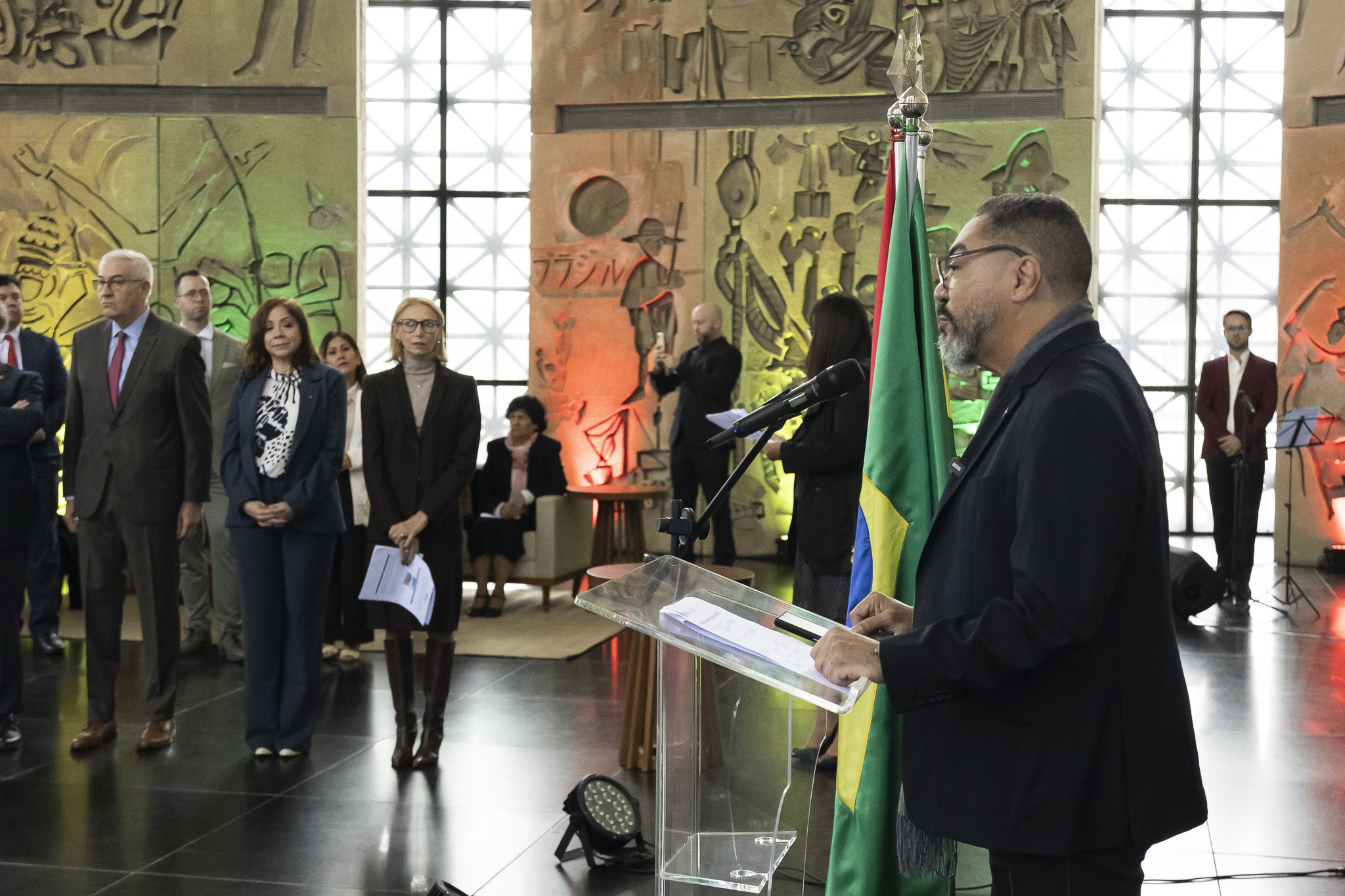 Participante pronuncia un discurso desde un atril transparente mientras el público sigue la ceremonia en un salón del Memorial
