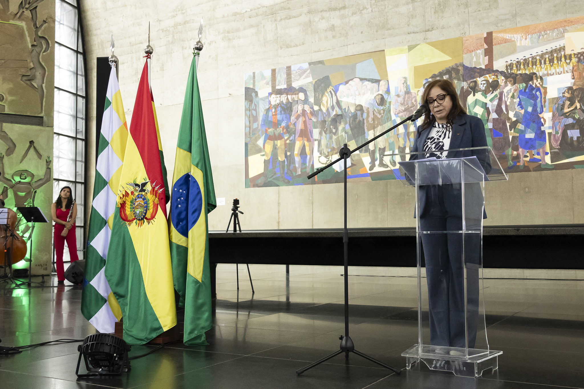 Vânia Claros pronuncia un discurso desde un atril transparente, ante las banderas de São Paulo, Bolivia y Brasil y un panel artístico en el Memorial