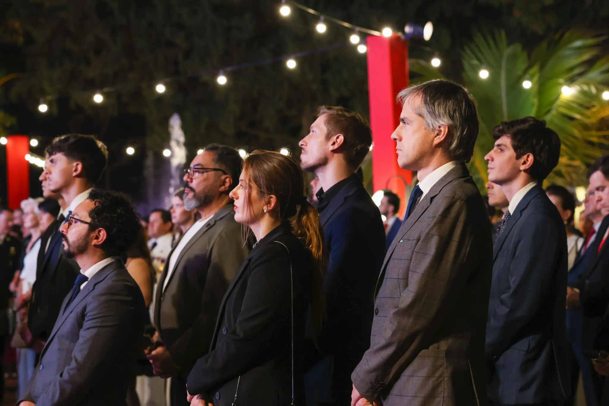 Representantes e invitados siguen de pie la ceremonia de las fiestas patrias de Chile en un espacio exterior iluminado