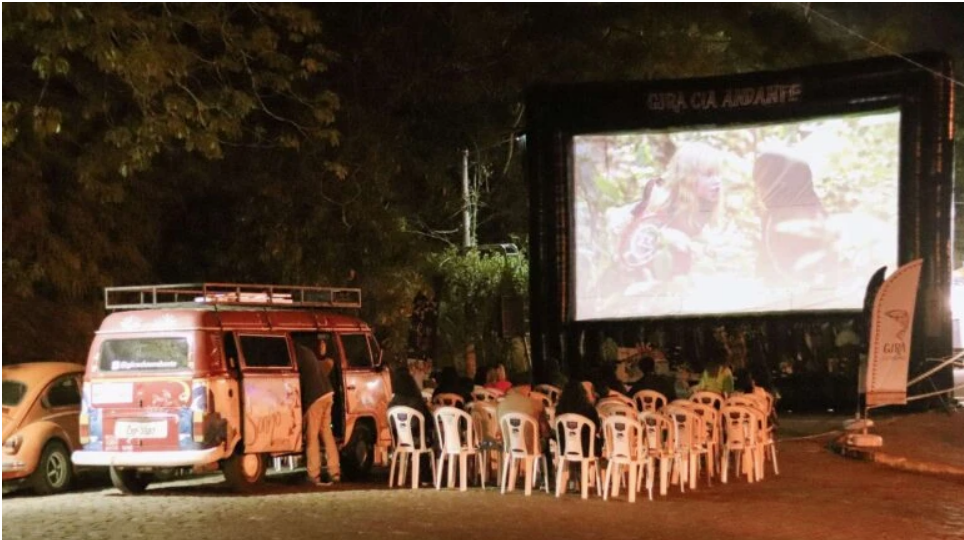 Referência visual de cinema itinerante noturno, com Kombi, tela de projeção e cadeiras organizadas ao ar livre