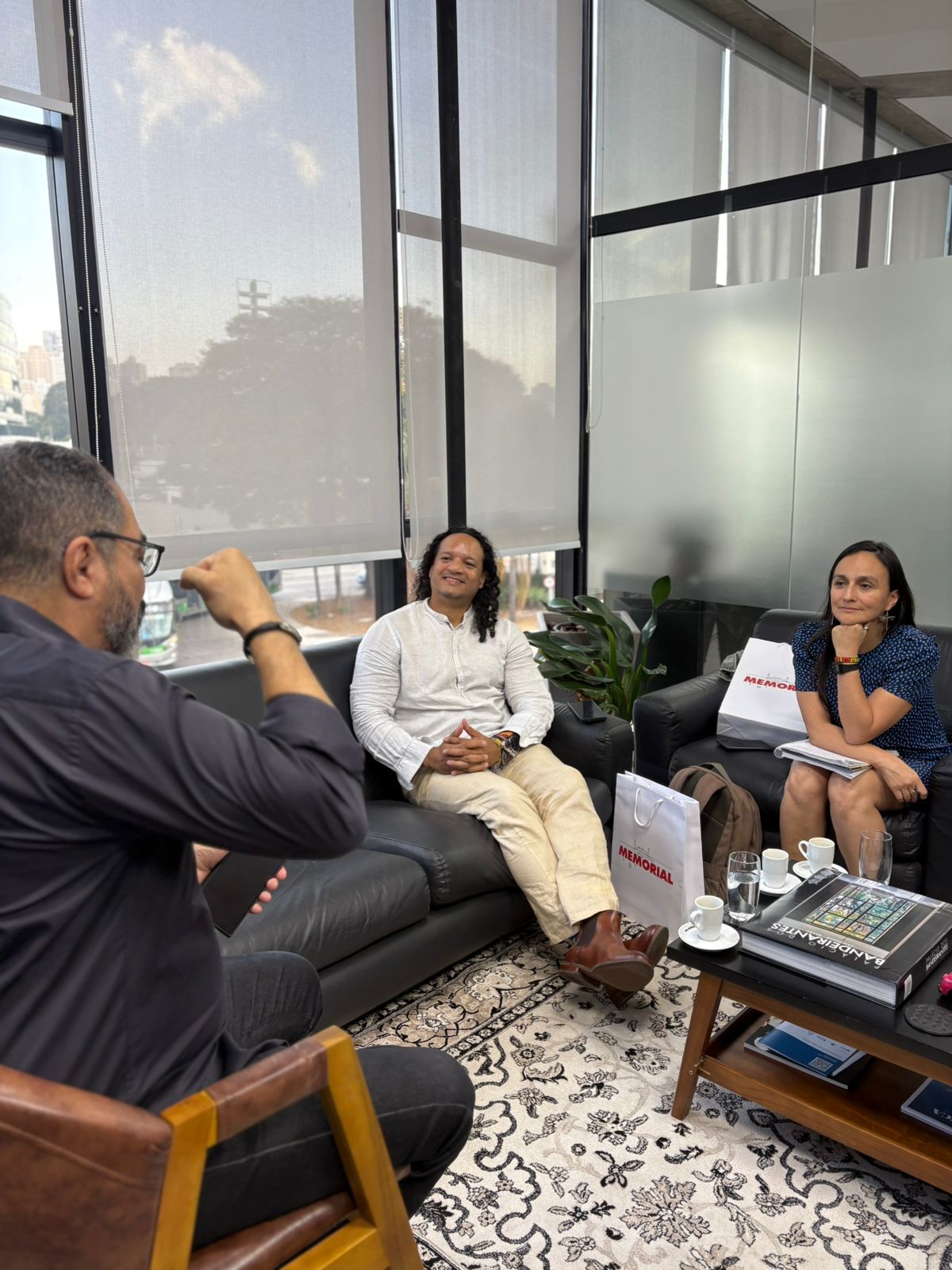 João Carlos Corrêa conversa com Carlos Arturo García e uma participante não identificada em reunião com livros, xícaras e materiais do Memorial