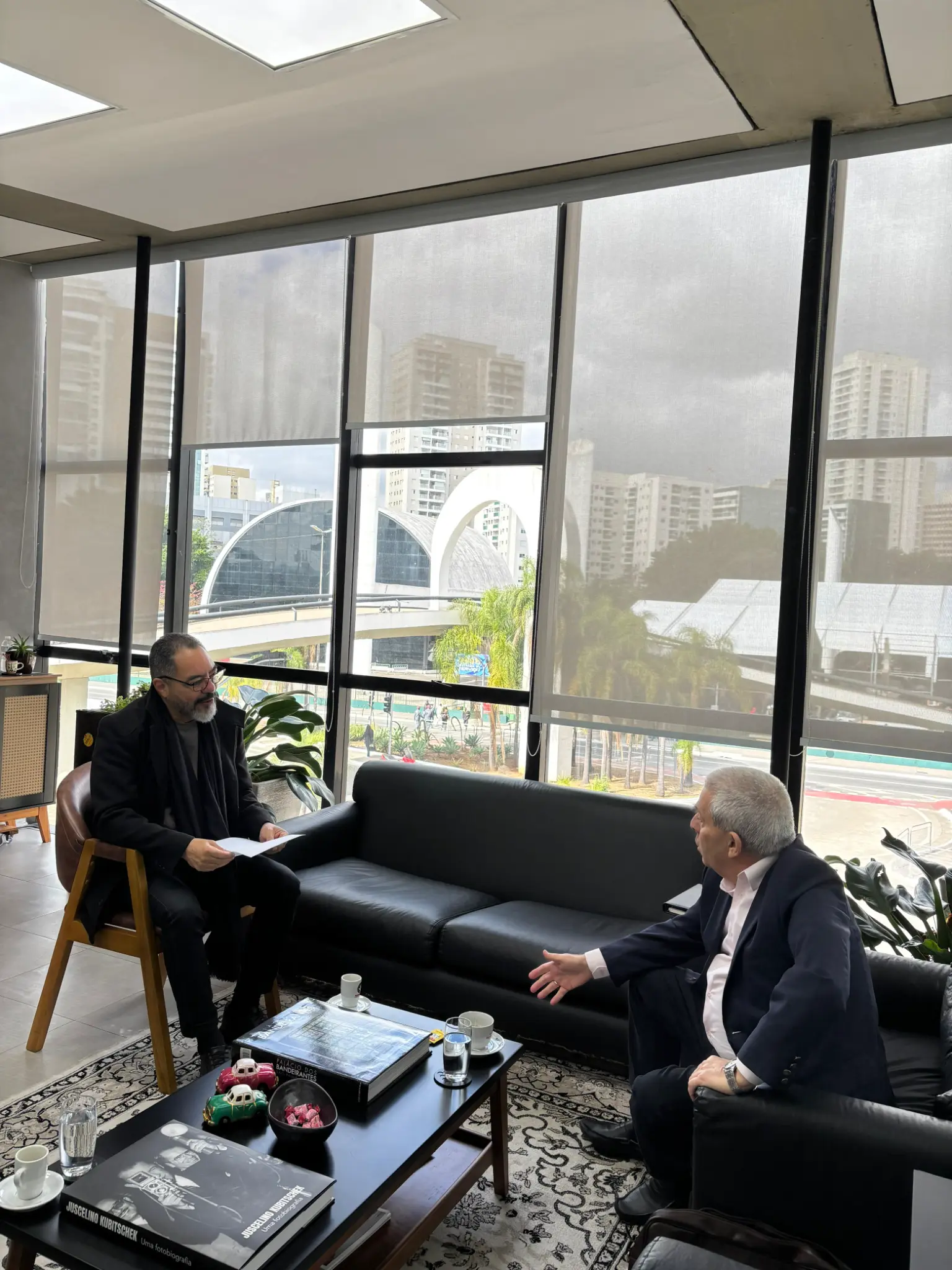 João Carlos Corrêa conversa con un representante sentado en un sillón, frente a una gran ventana con vista al Memorial y a la ciudad