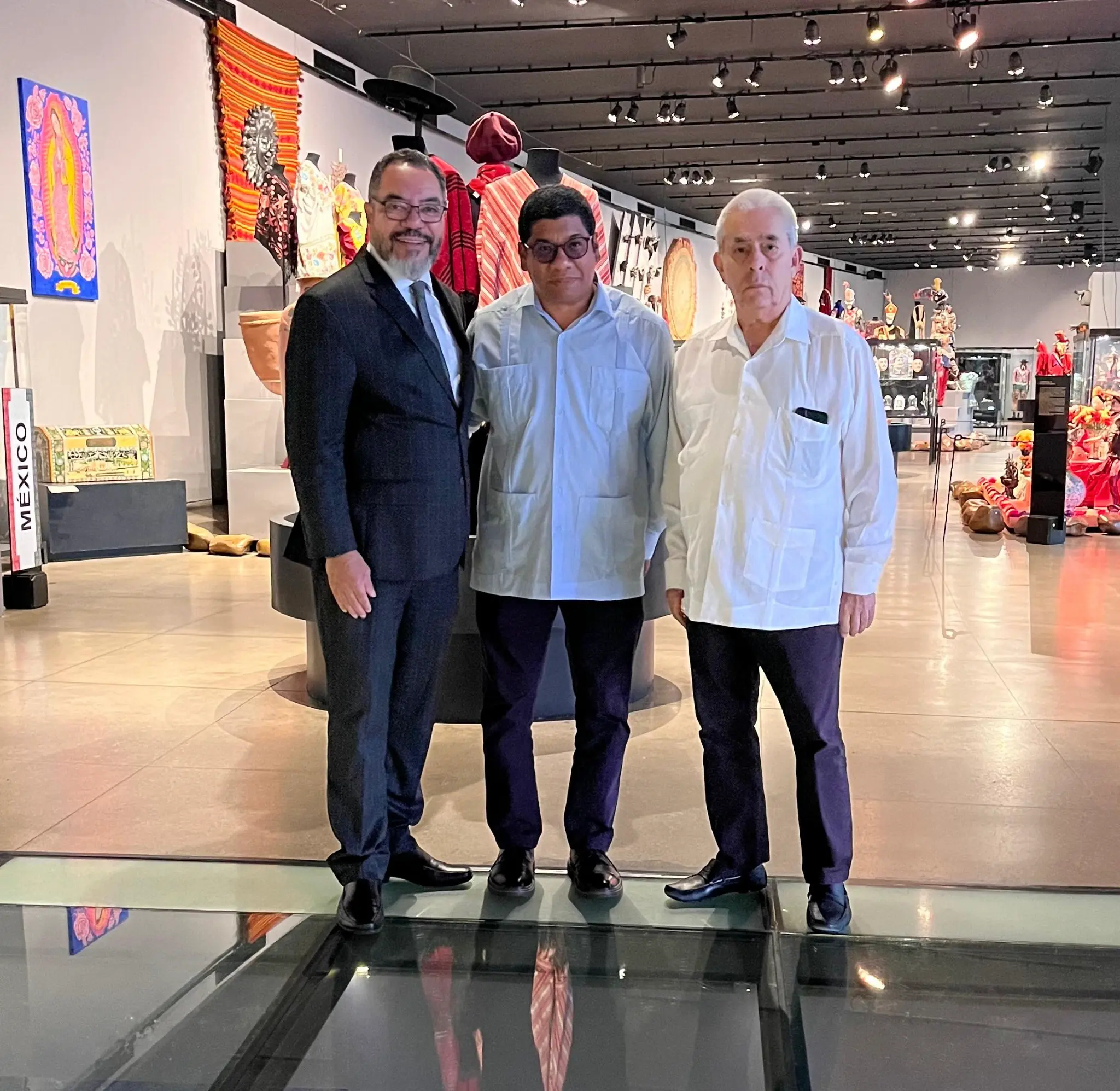 João Carlos Corrêa y dos participantes posan en una sala de exposiciones con vestimentas, objetos y piezas de artesanía latinoamericana