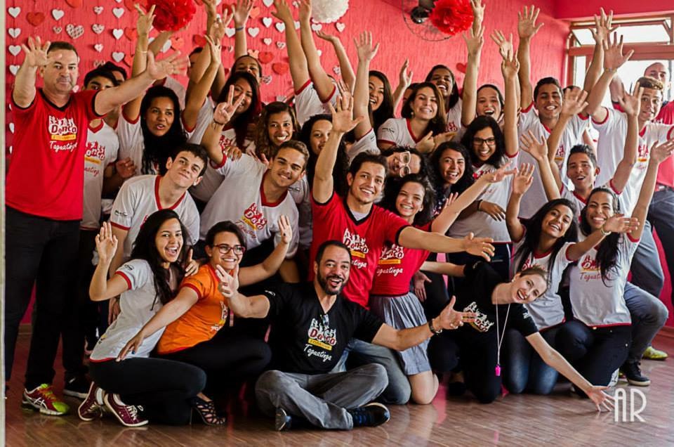 Grupo numeroso de participantes com figurinos variados dança em área externa coberta