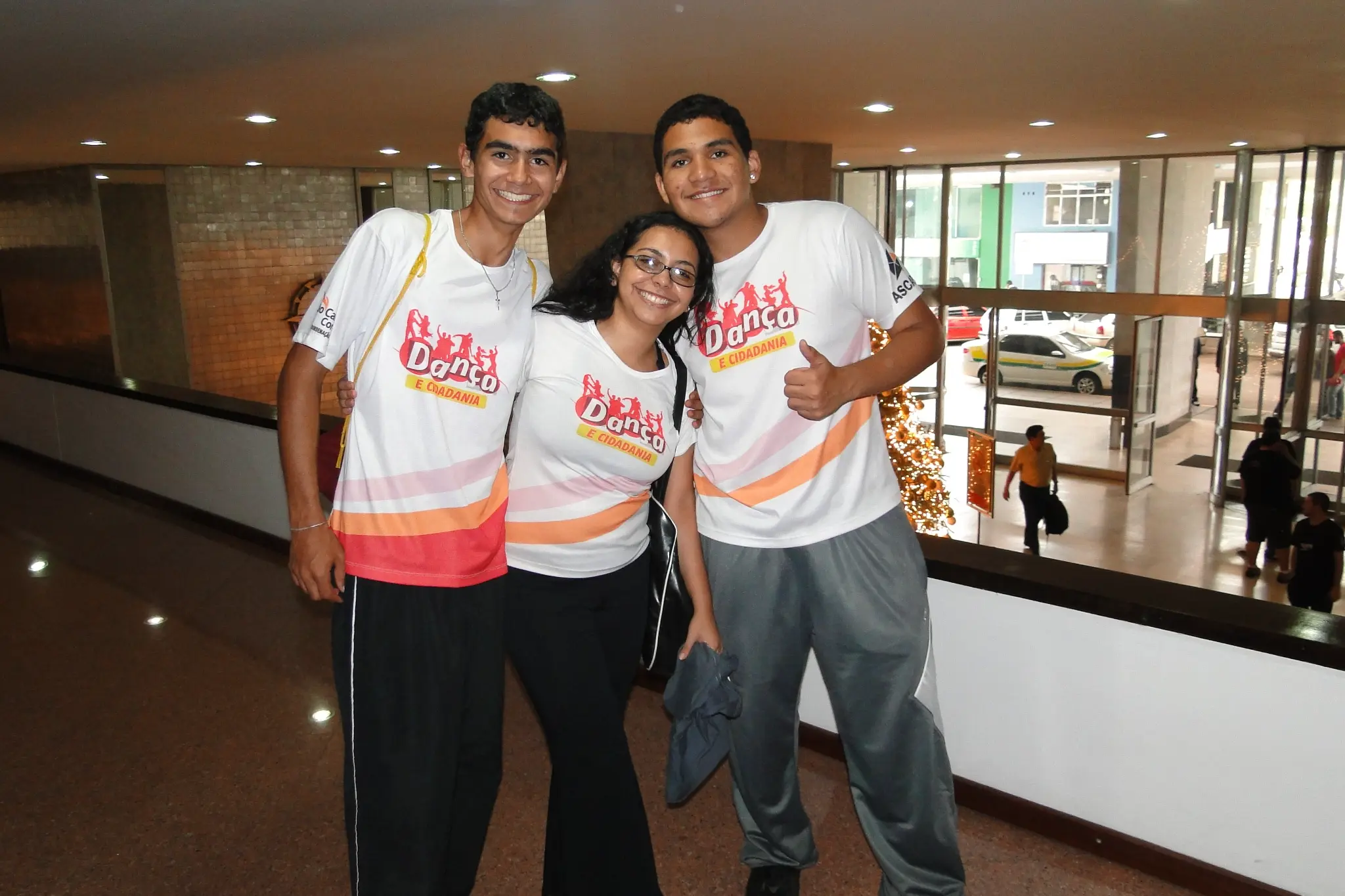 Três participantes com camisetas do Projeto Dança e Cidadania reunidos em uma área interna