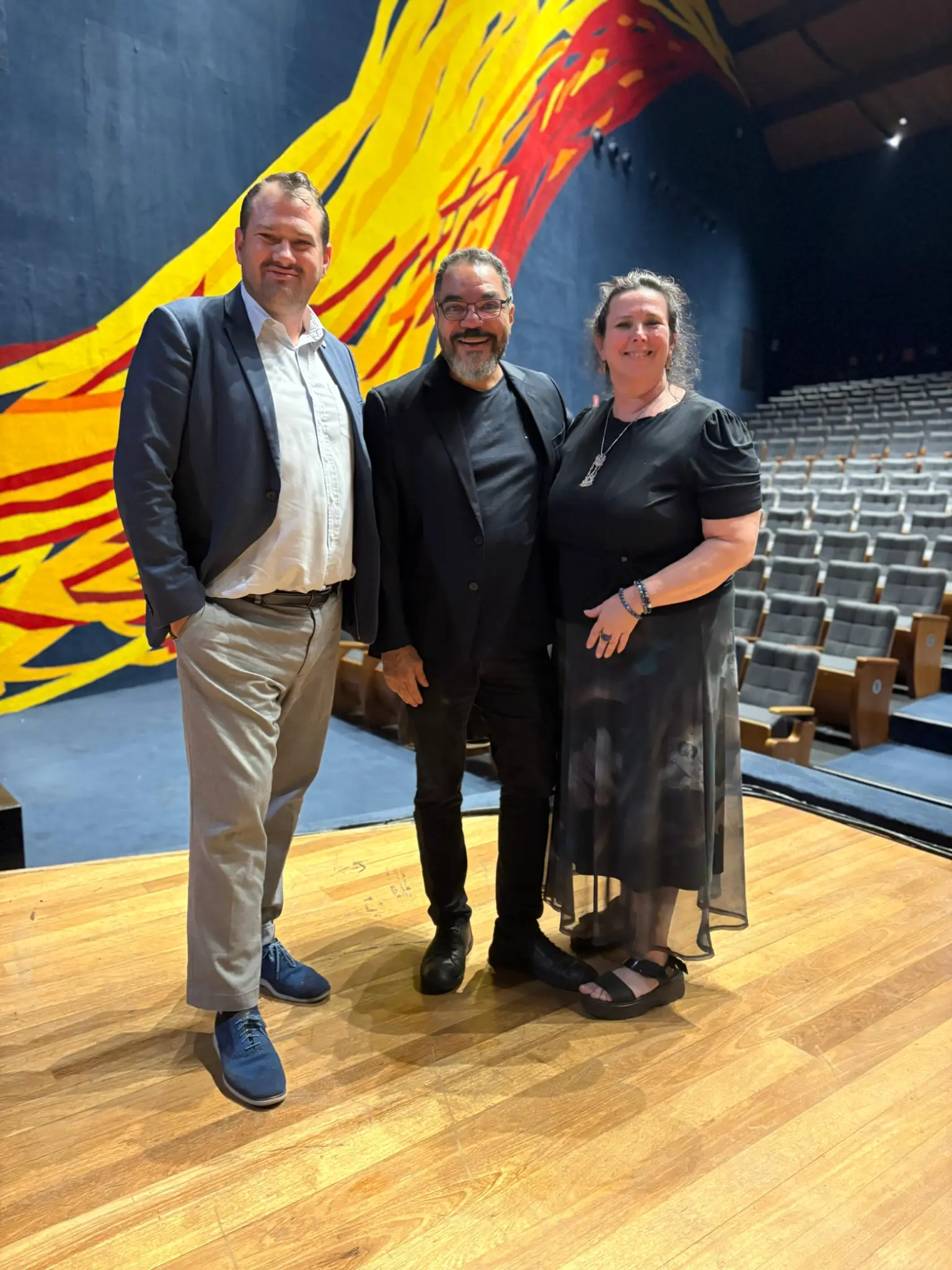 Gavin Nardocchio-Jones, João Carlos Corrêa y Dina E. Thrasher posan en el escenario del Auditório Simón Bolívar frente a un gran panel colorido