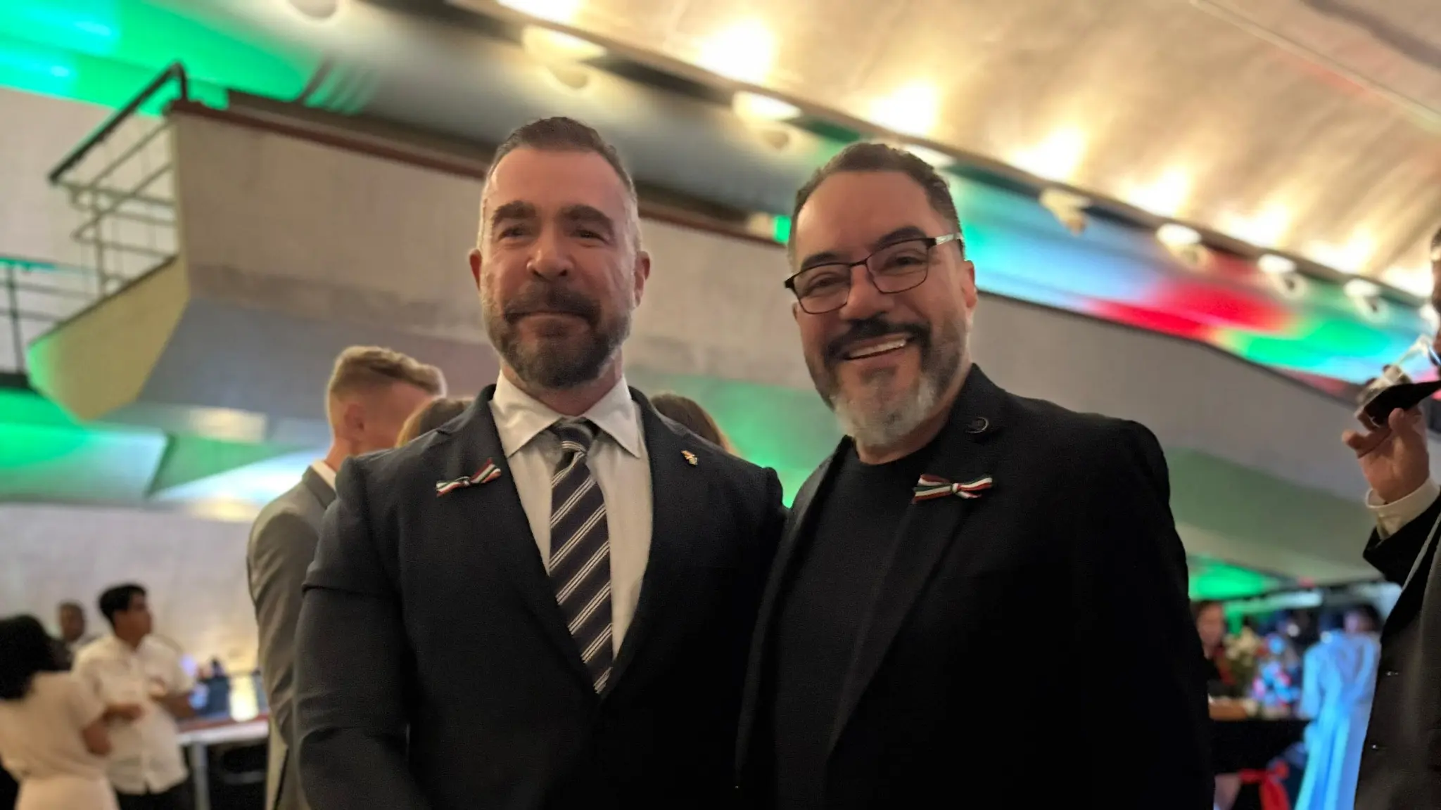 Daniel Gallego Arcas y João Carlos Corrêa posan uno al lado del otro en un evento cultural, con iluminación verde y roja al fondo