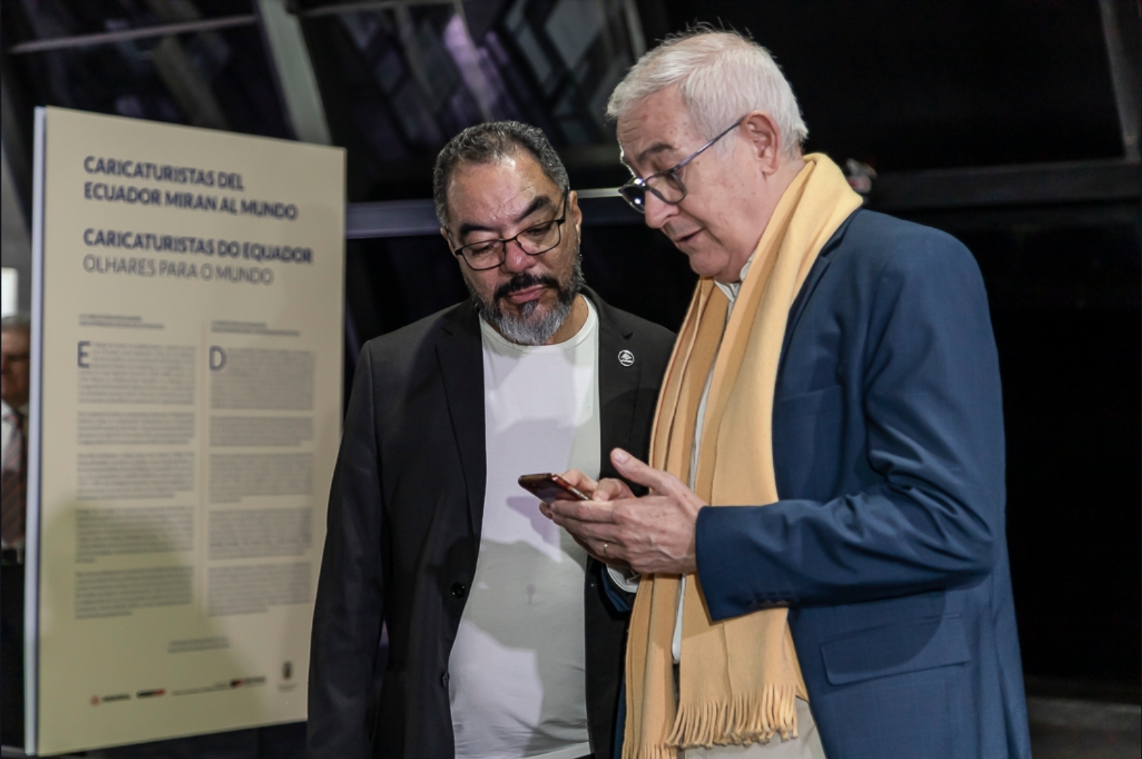 João Carlos Corrêa y Luis Fernando Ávalos Giménez observan juntos un teléfono celular frente al panel de la exposición Caricaturistas do Equador