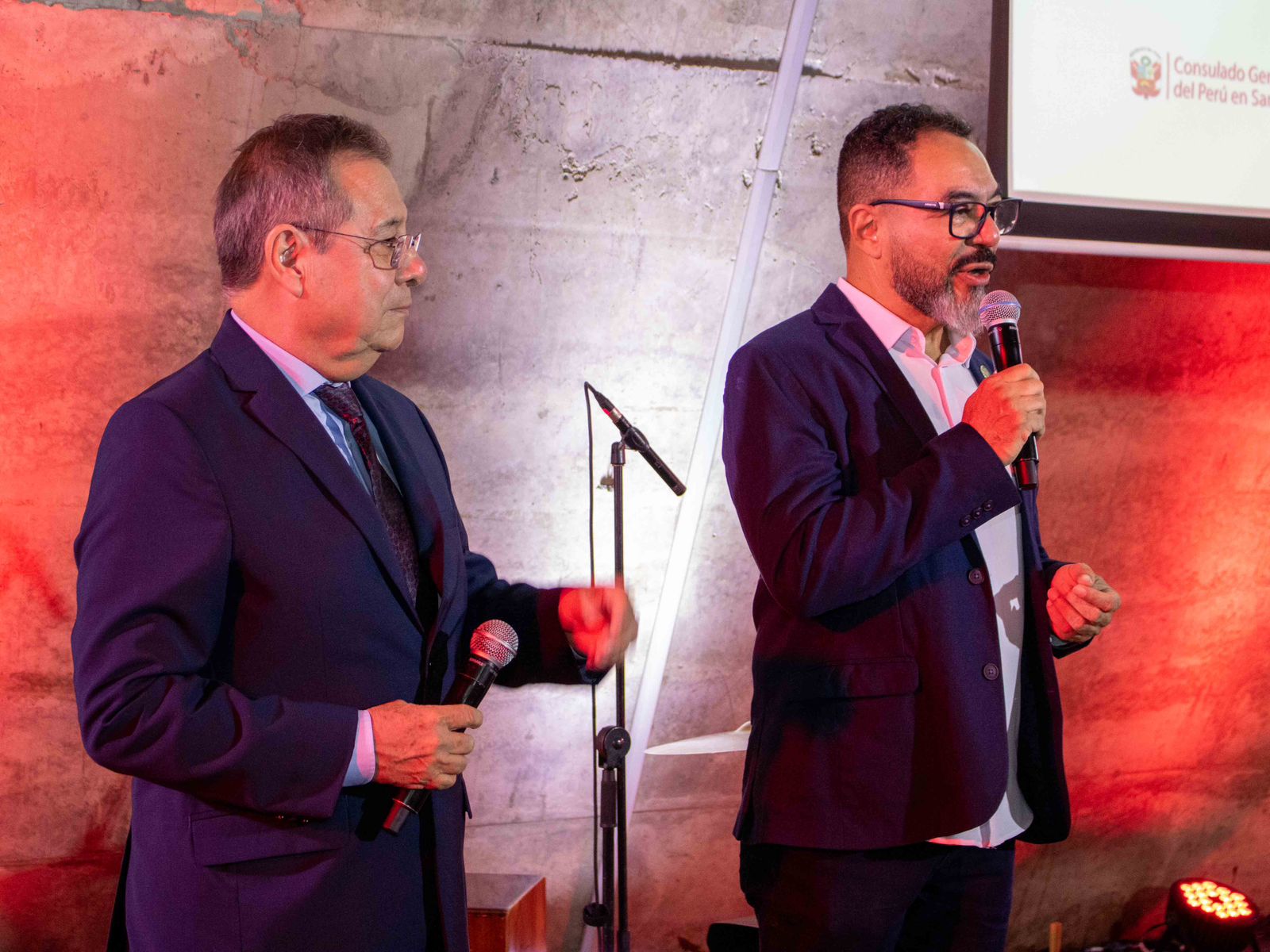 Luis Armando Monteagudo Pacheco acompaña a João Carlos Corrêa, quien habla al micrófono en un escenario con iluminación roja y una proyección del consulado peruano