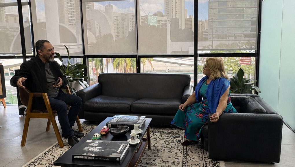 João Carlos Corrêa y Hanoi Sánchez Paniagua conversan sentados en un entorno de trabajo, con una mesa, libros y ventanas al fondo