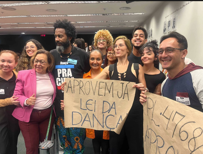 Grupo de mobilização segura cartazes com pedidos pela aprovação da Lei da Dança em espaço legislativo.