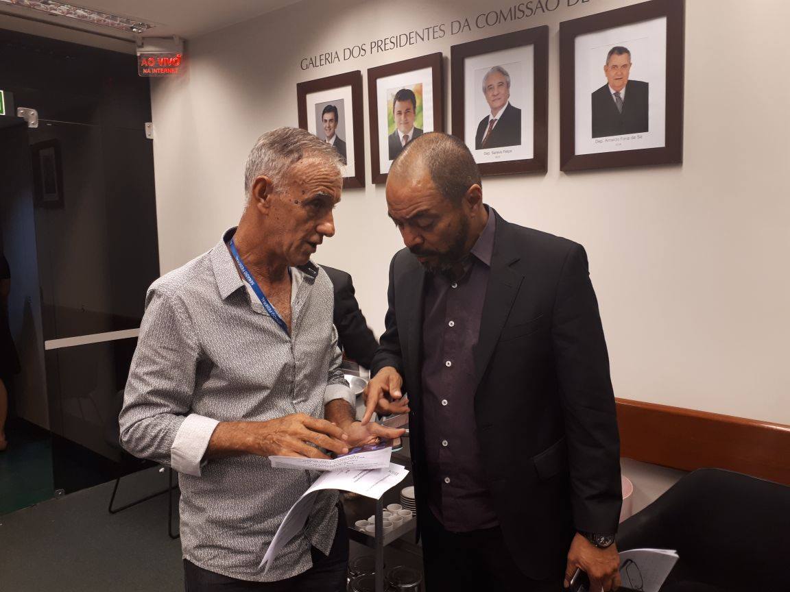 Participantes acompanham atividade em auditório com mesas, documentos e microfones.