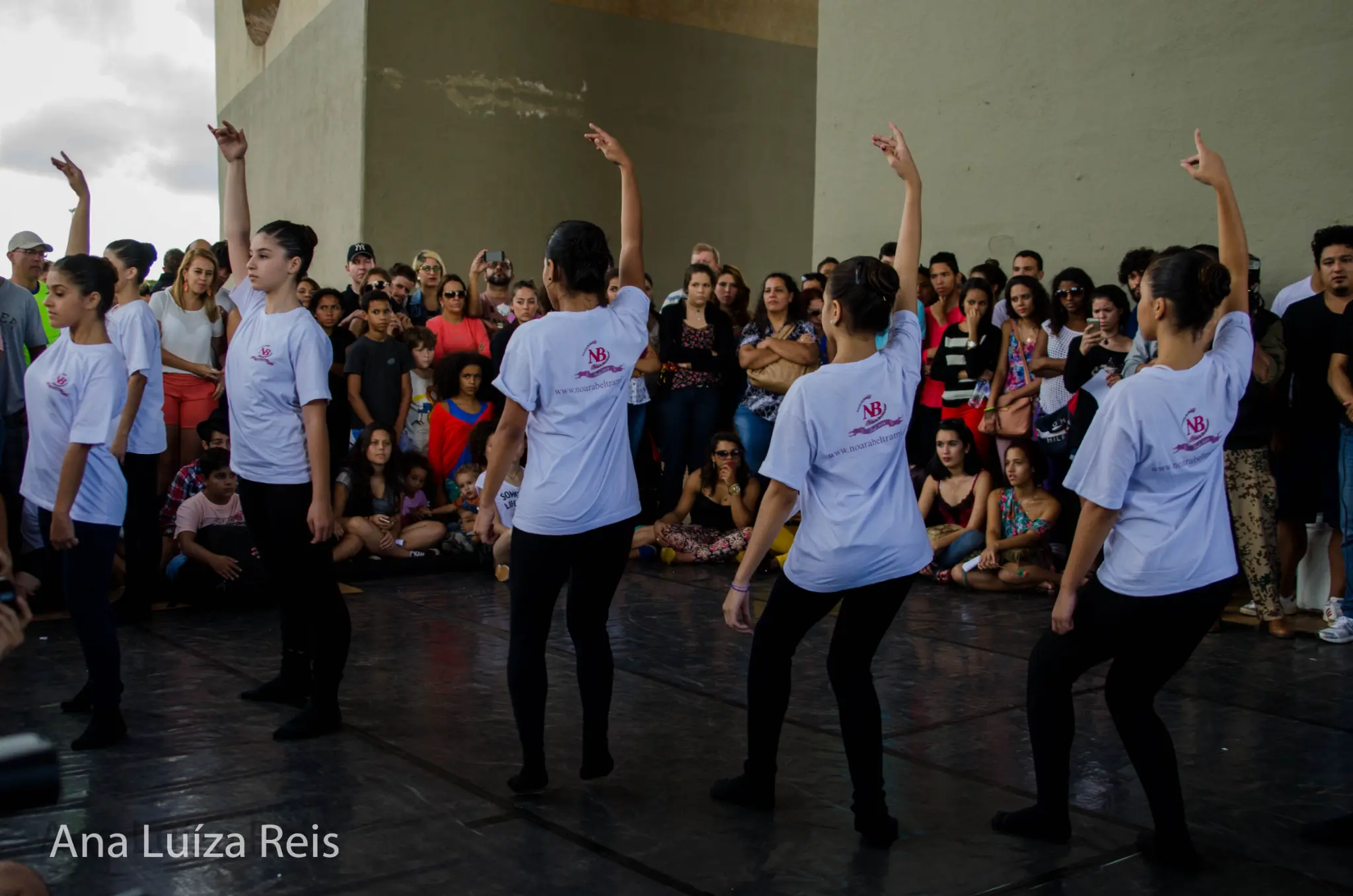 Grupo de jovens em camisetas brancas e calças pretas apresenta uma coreografia diante do público.