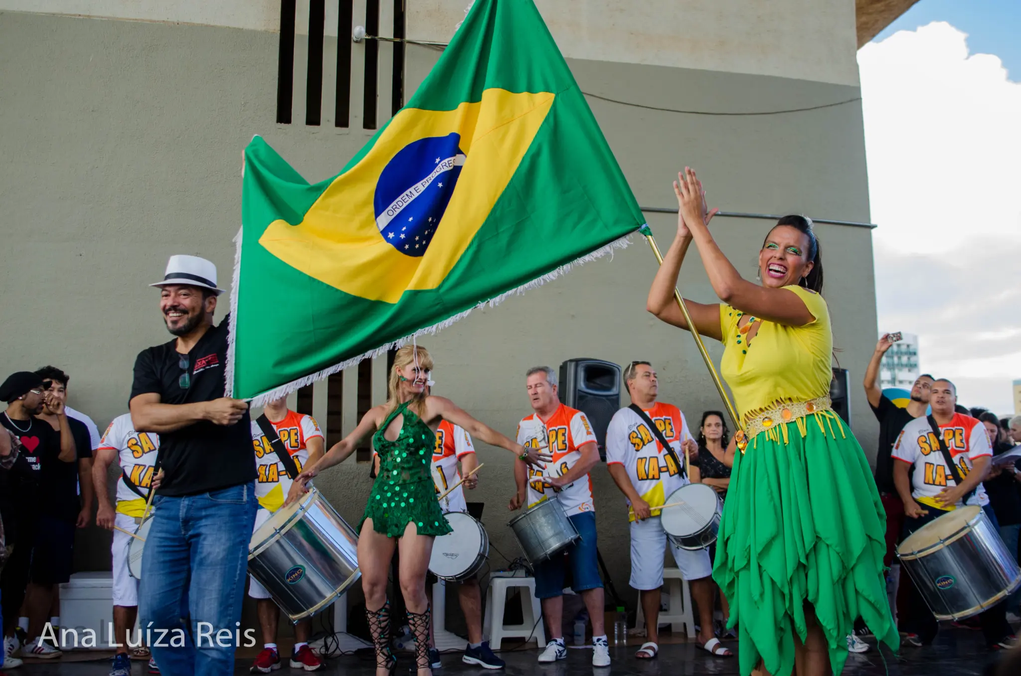 Participantes sorriem com a bandeira do Brasil enquanto músicos aparecem ao fundo.