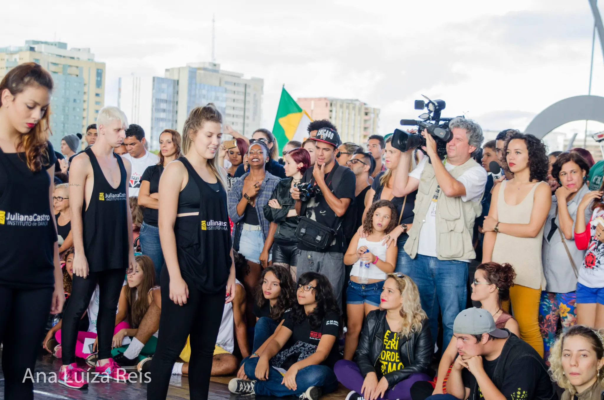 Público e participantes acompanham uma atividade ao ar livre enquanto um cinegrafista faz o registro.