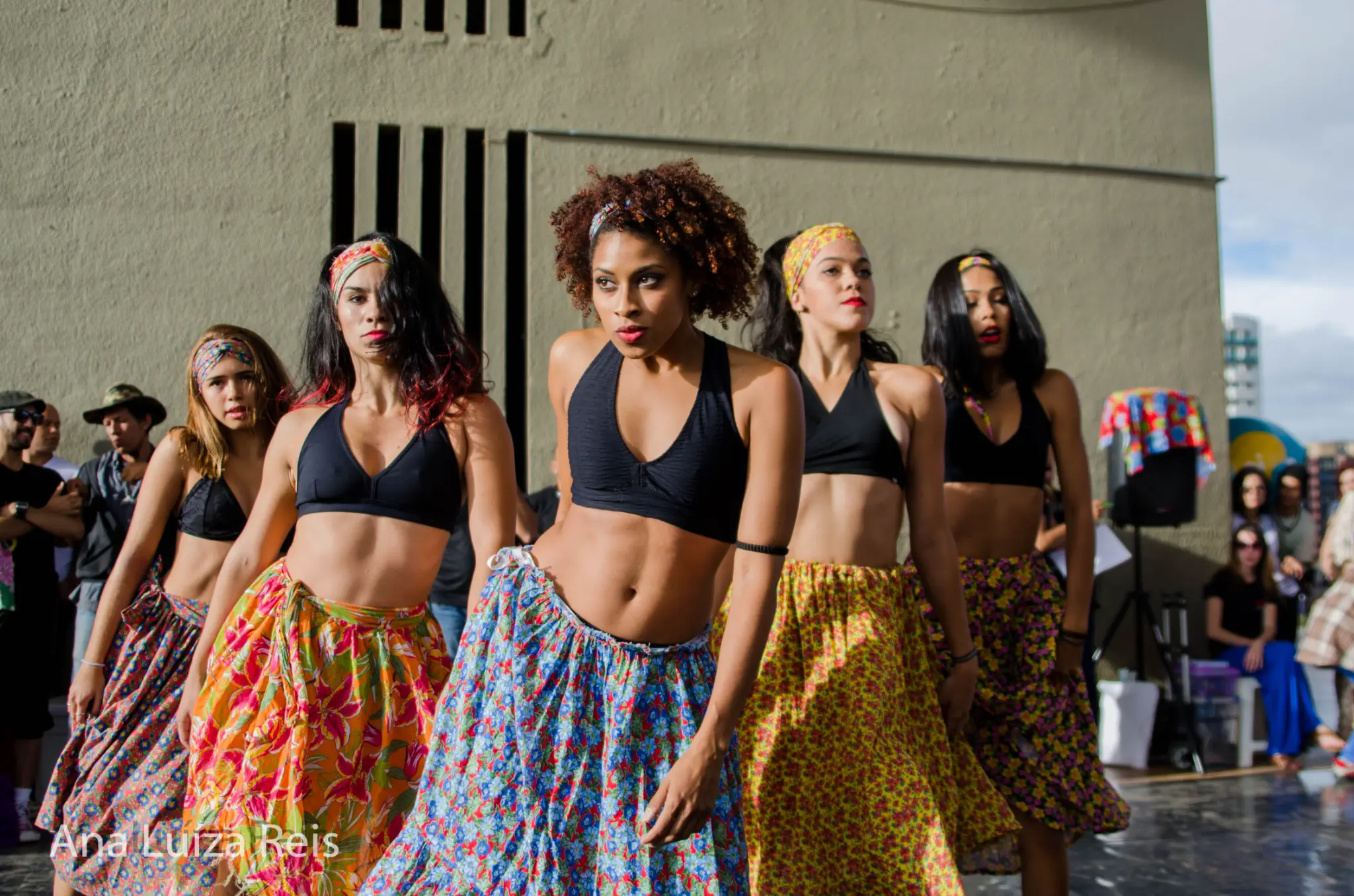 Grupo de mulheres com saias longas estampadas dança em formação ao ar livre.