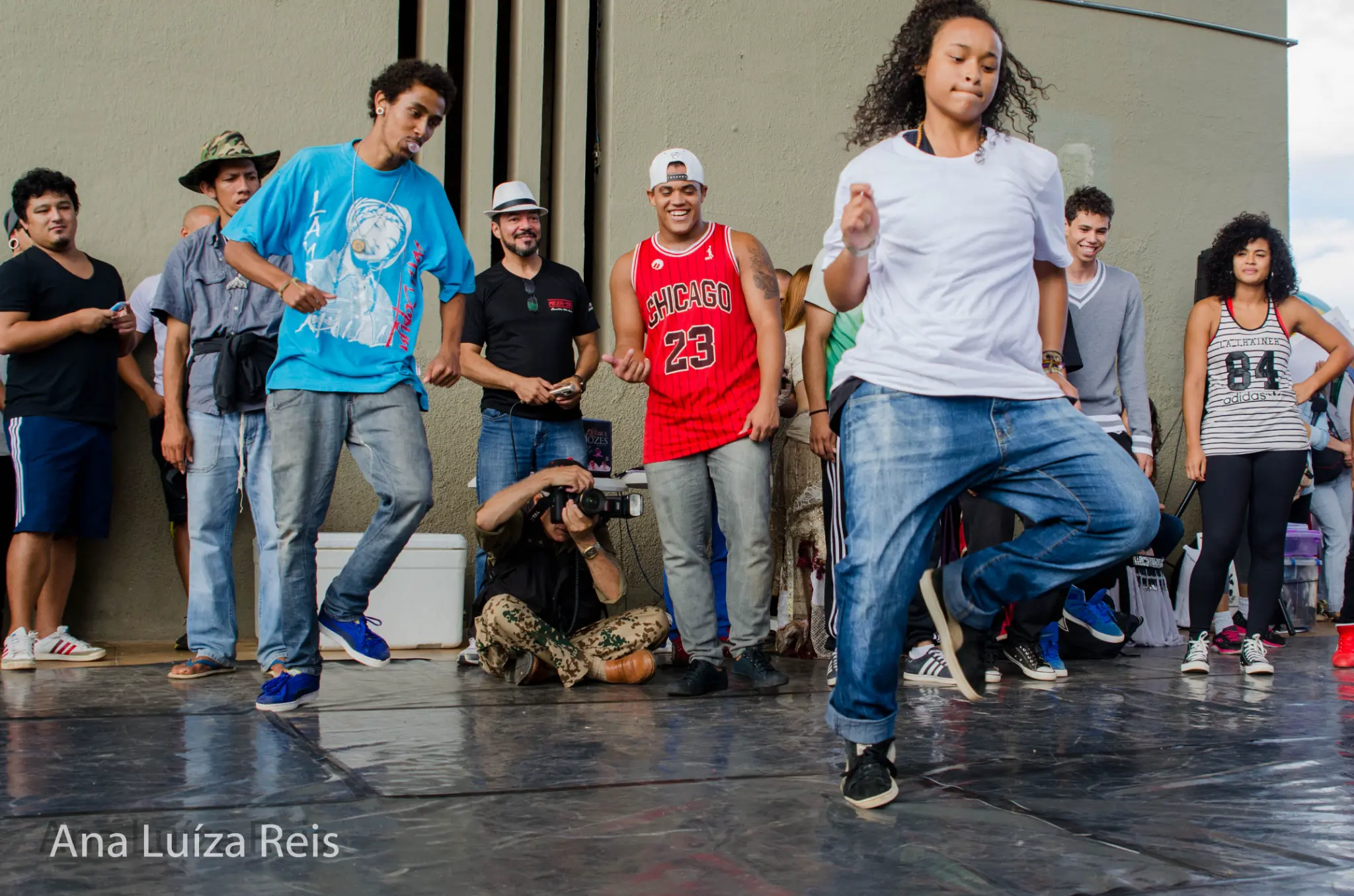 Dois jovens executam movimentos de dança urbana enquanto espectadores e um fotógrafo acompanham a apresentação.