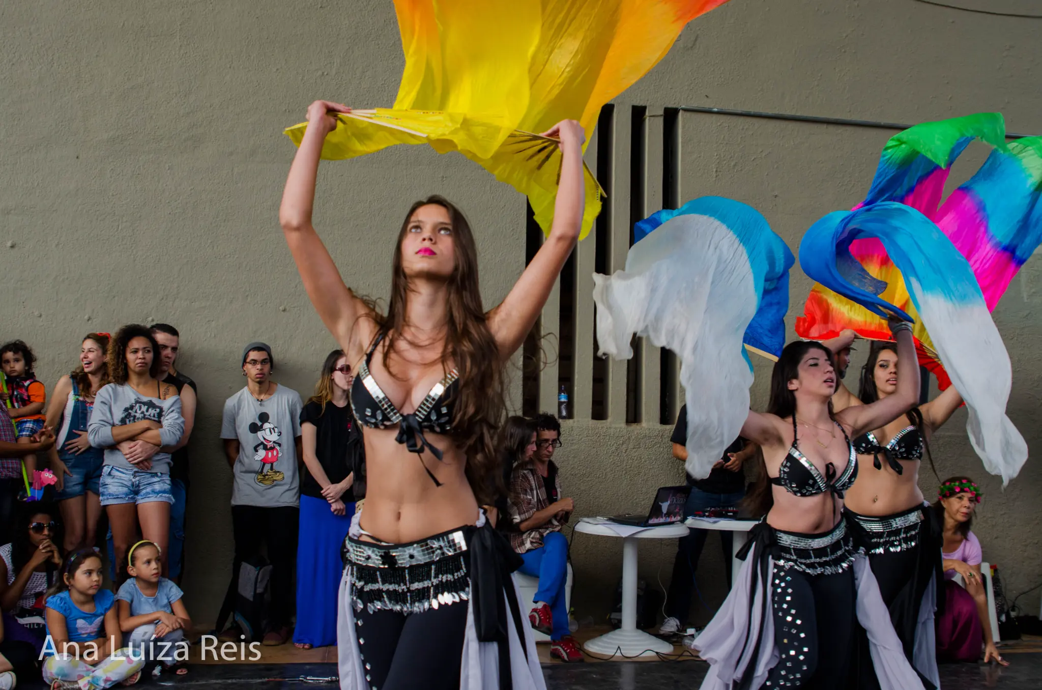 Dançarinas com trajes escuros movimentam véus coloridos diante dos espectadores.