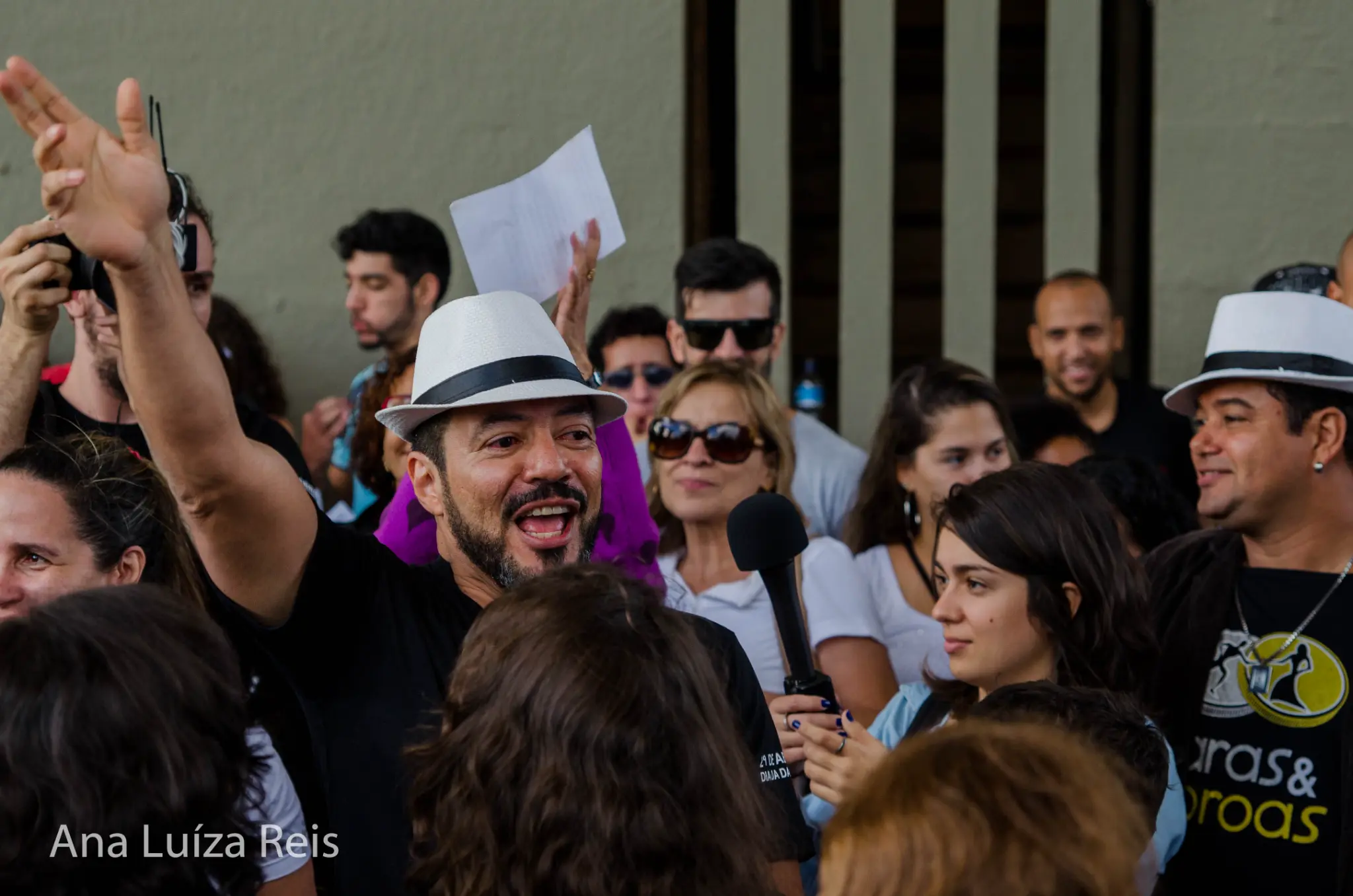 Participante com chapéu branco e braço erguido fala ao microfone em meio ao público reunido ao ar livre.