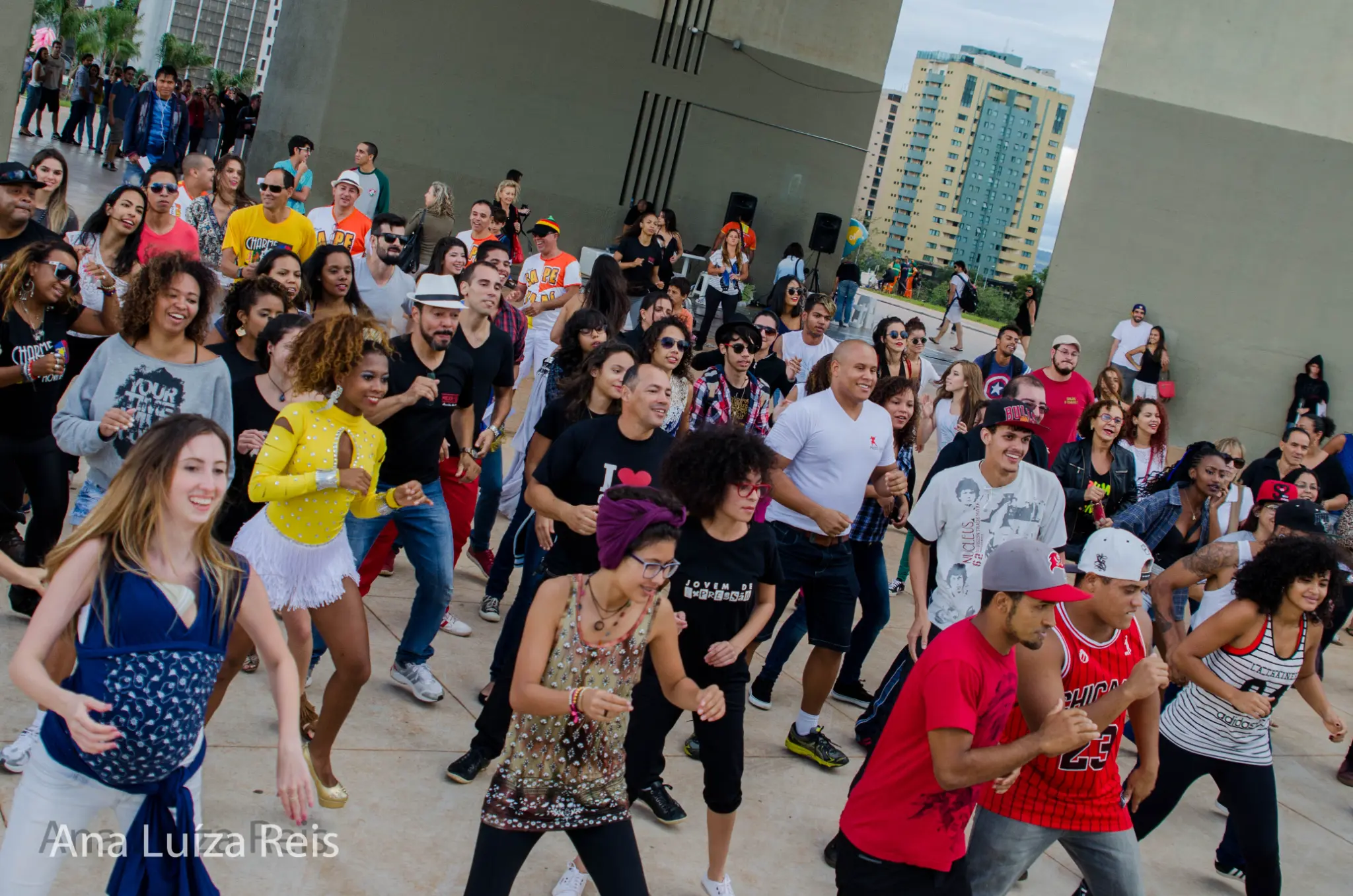 Grande grupo de pessoas participa de uma coreografia coletiva em espaço público durante o dia.
