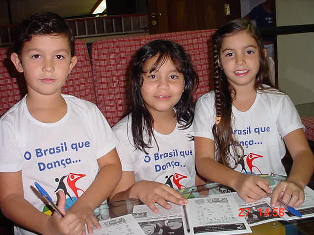 Três crianças com camisetas da I Maratona de Dança reunidas junto a materiais impressos