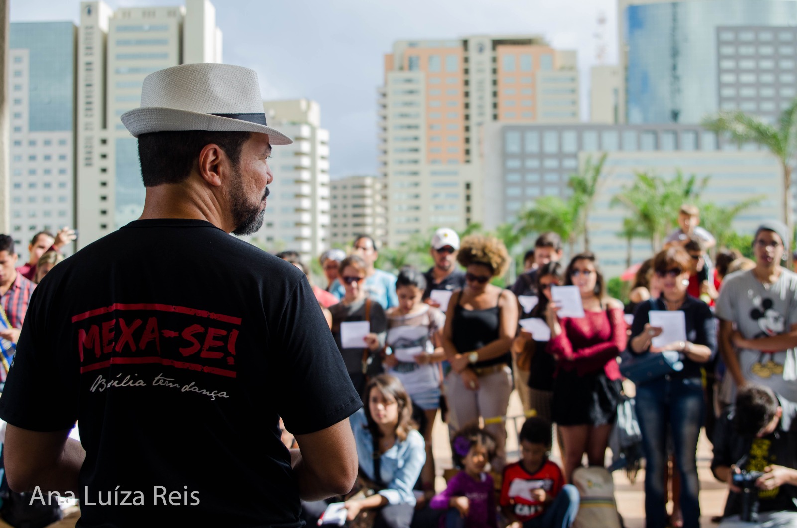 João Carlos Corrêa de costas, usando camiseta do projeto MEXA-SE! Brasília tem dança, diante de participantes em espaço público; crédito visível: Ana Luíza Reis