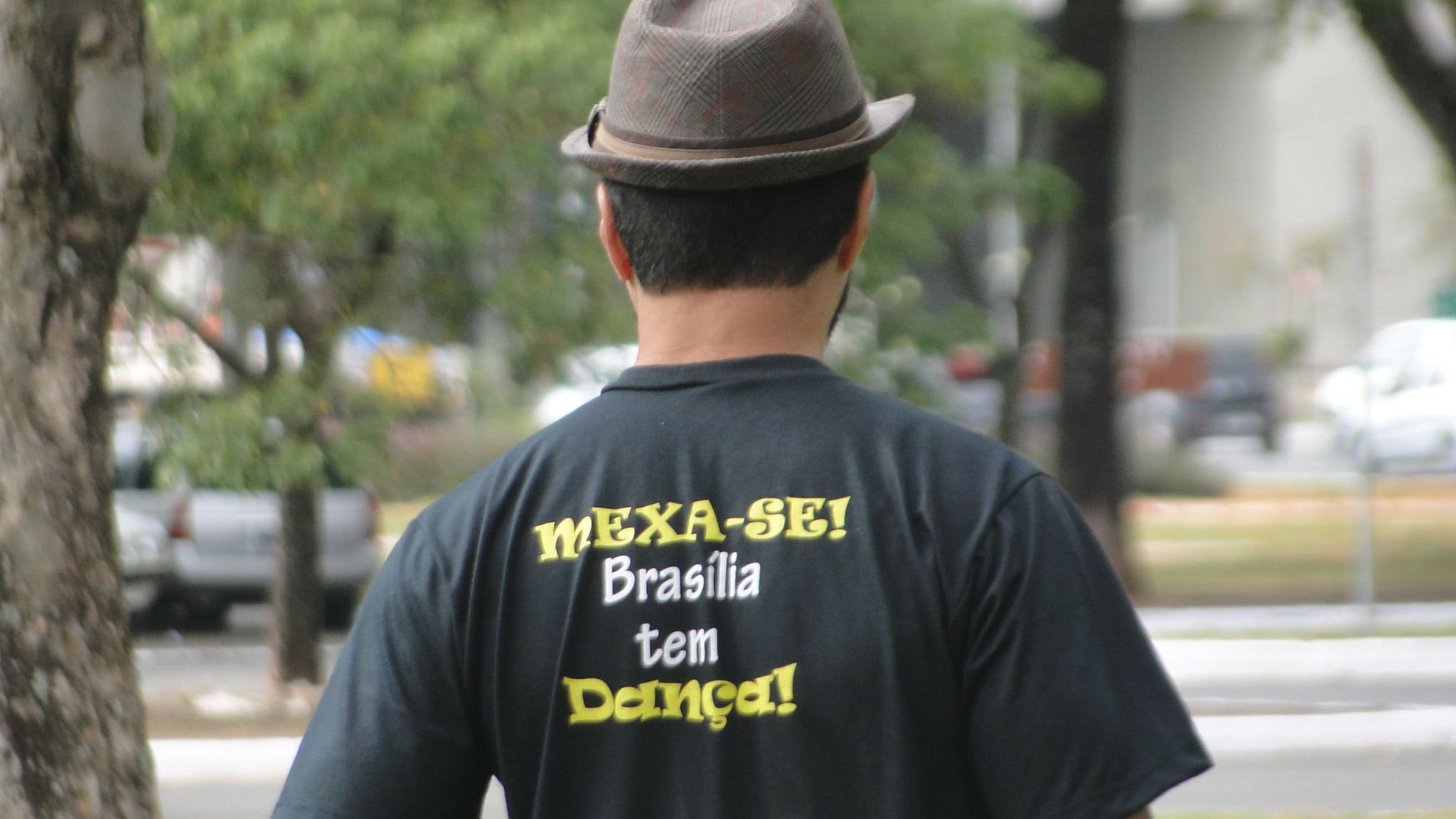 Pessoa de costas usa chapéu e camiseta preta com a inscrição MEXA-SE! Brasília tem Dança em uma área externa arborizada.