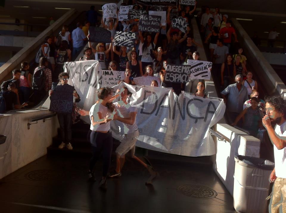 Manifestantes ocupam uma escadaria com faixas e cartazes enquanto duas pessoas dançam no primeiro plano.