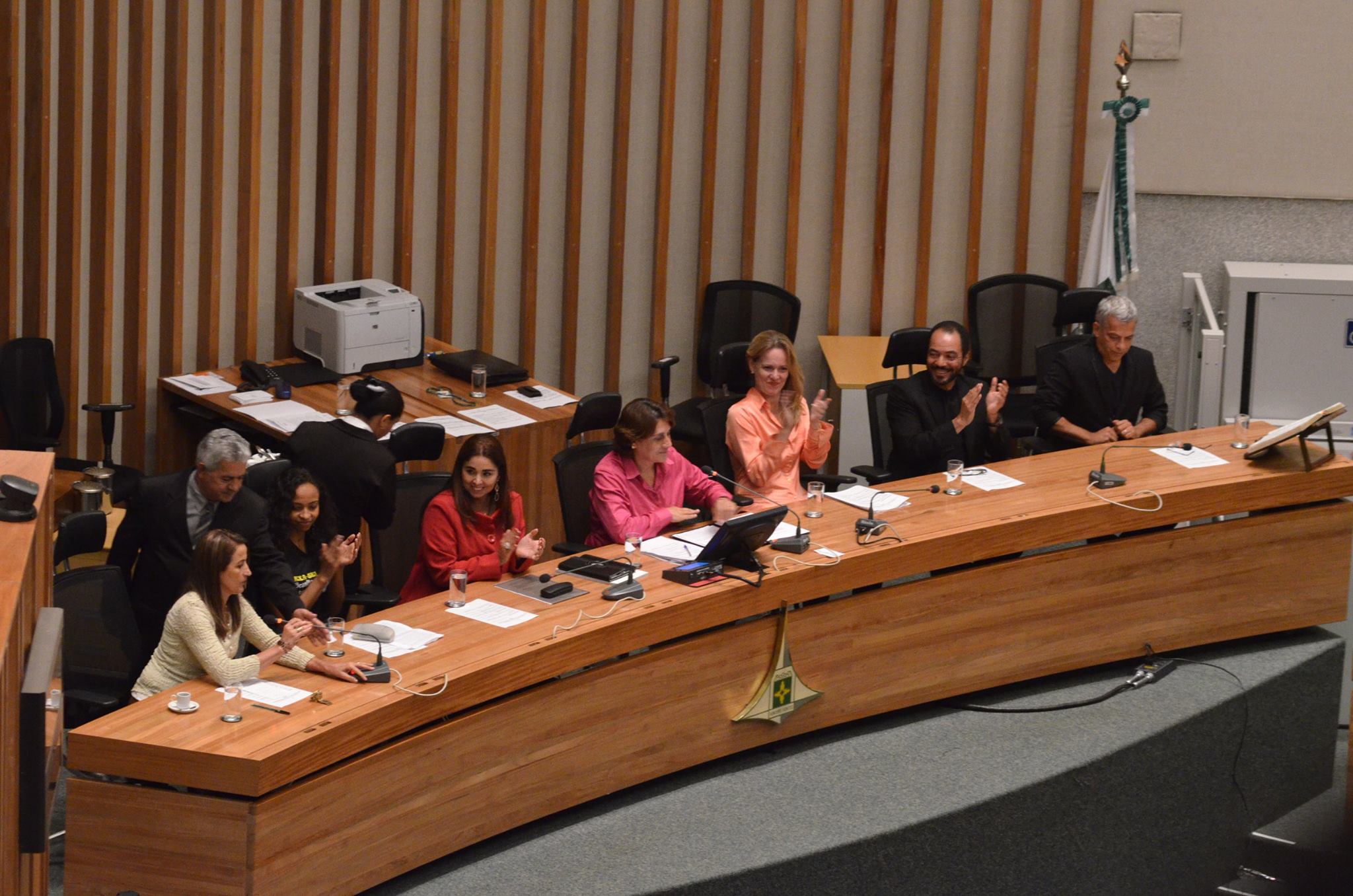 Vista ampla de participantes reunidos em uma mesa institucional equipada com microfones e documentos.