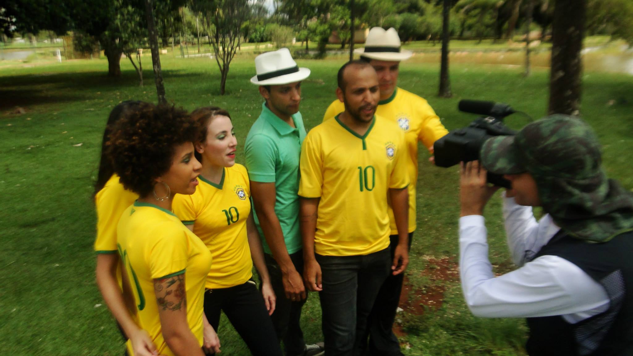 Grupo com roupas nas cores verde e amarela participa de gravação audiovisual em uma área verde.