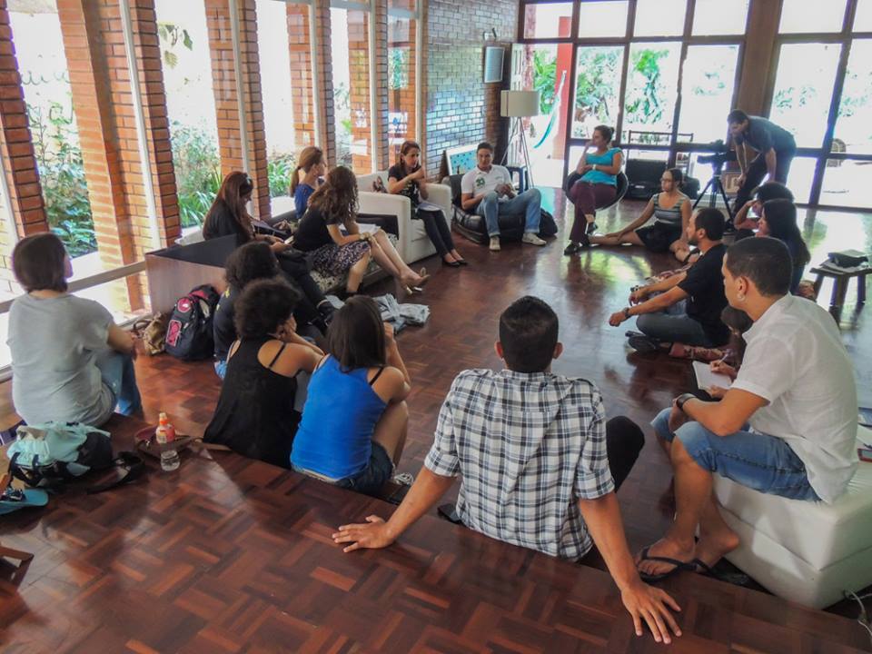 Grupo de pessoas reunido em círculo em uma sala ampla, com participantes sentados em sofás e no piso.