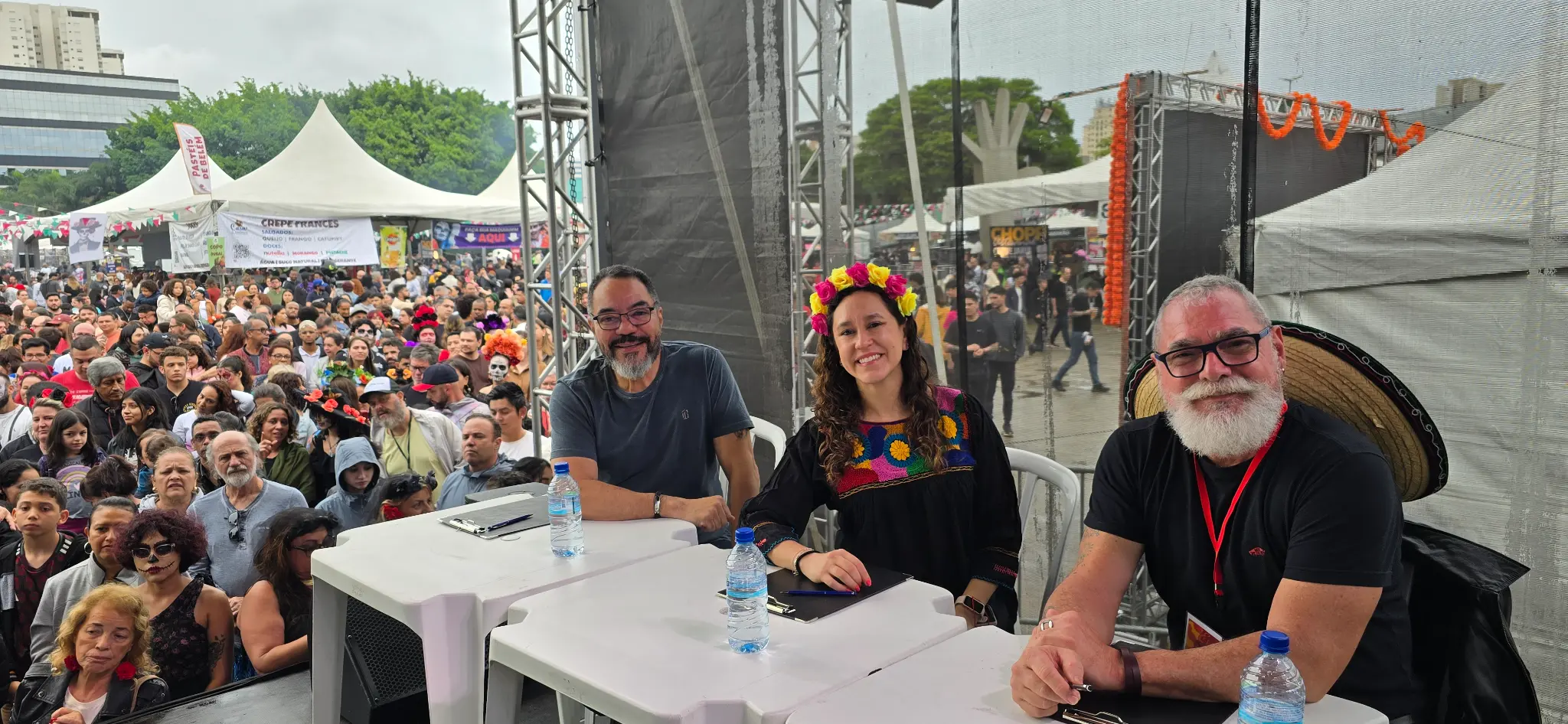 Três jurados sentados atrás de mesas com pranchetas e público reunido ao fundo